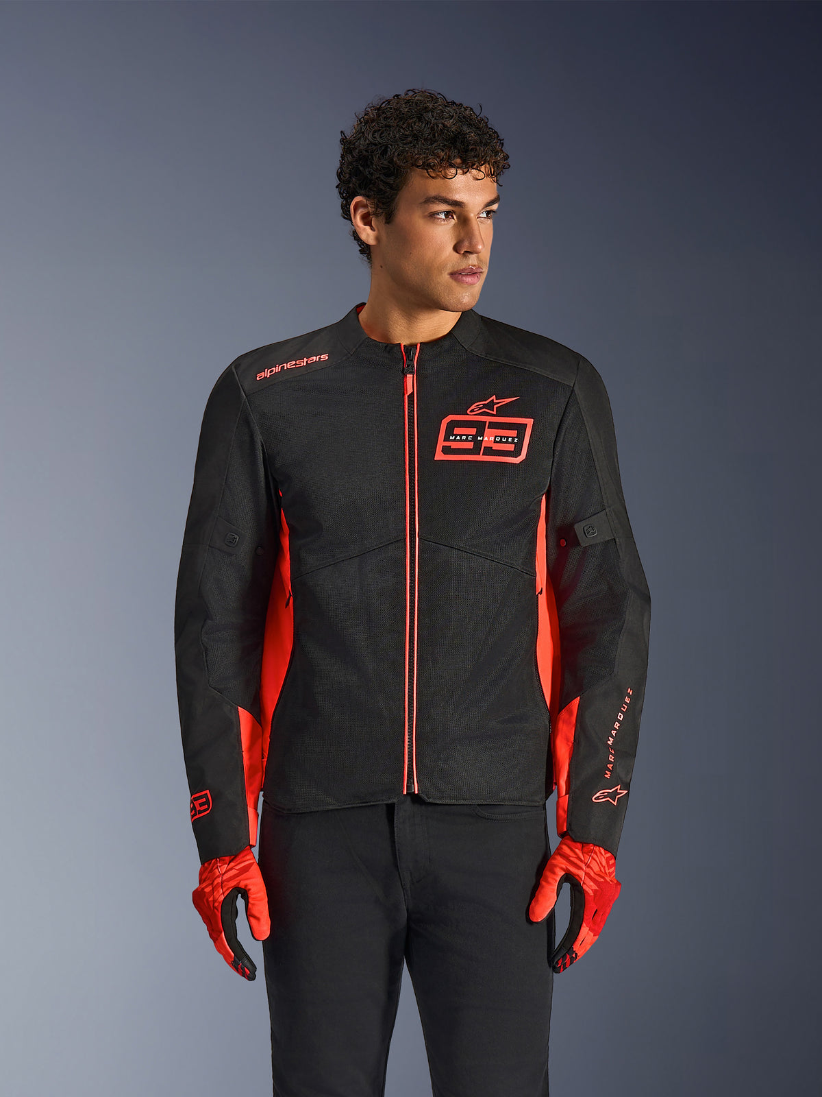 Giacca Alpinestars MM93 C-1 Air, giacca da moto urbana, rosso fluo e nero, indossata da un modello con guanti rossi abbinati e pantaloni neri, con il logo 93 Marc Marquez sul petto e pannelli in rete per il flusso d'aria