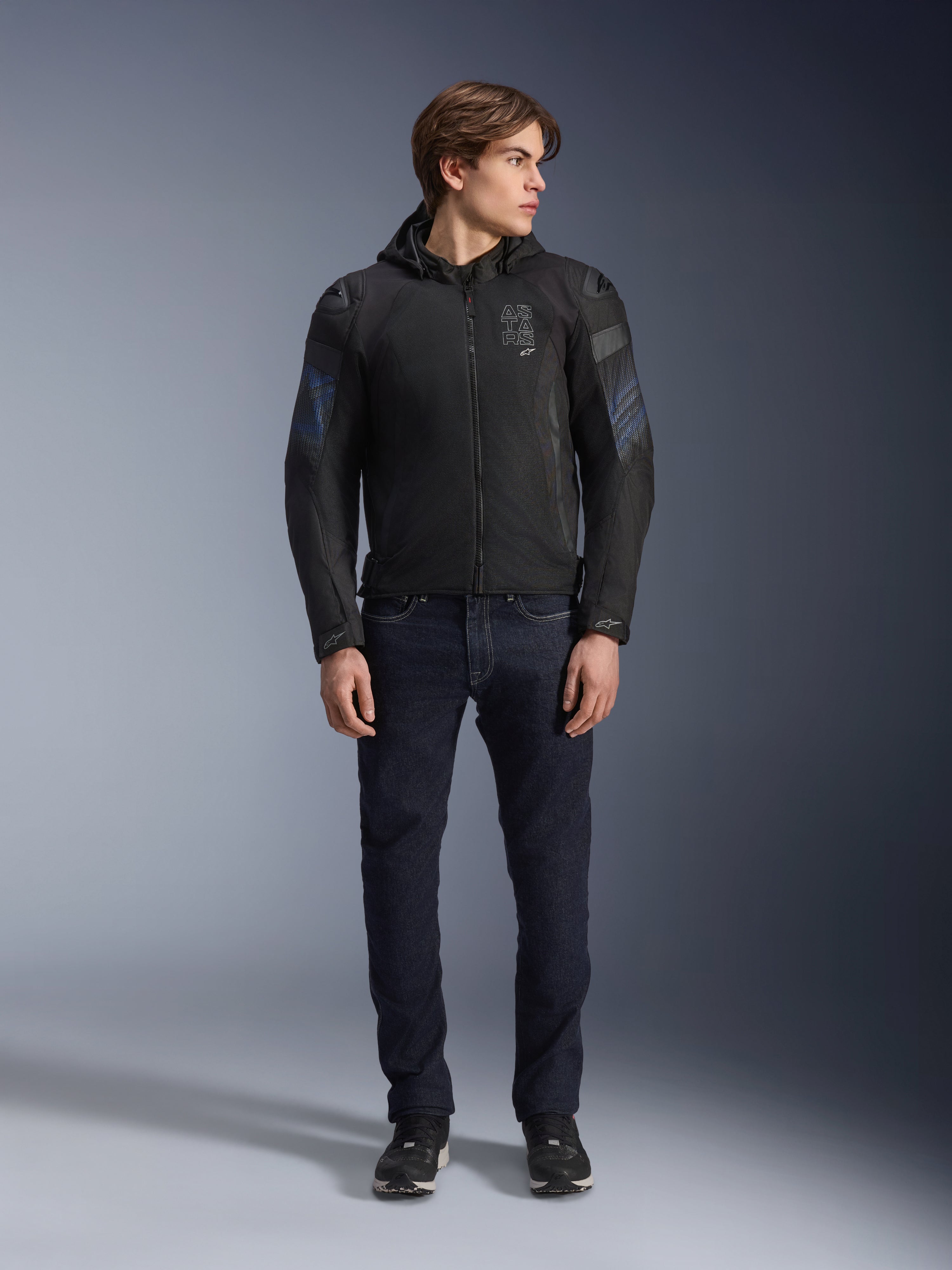 Modello che indossa la giacca Alpinestars Zaca Air Venom WP in nero e blu elettrico, giacca da moto in tessuto con pannelli in rete 3D e cappuccio rimovibile, abbinata a jeans in denim scuro, abbigliamento tecnico da guida di ispirazione sportiva moderna