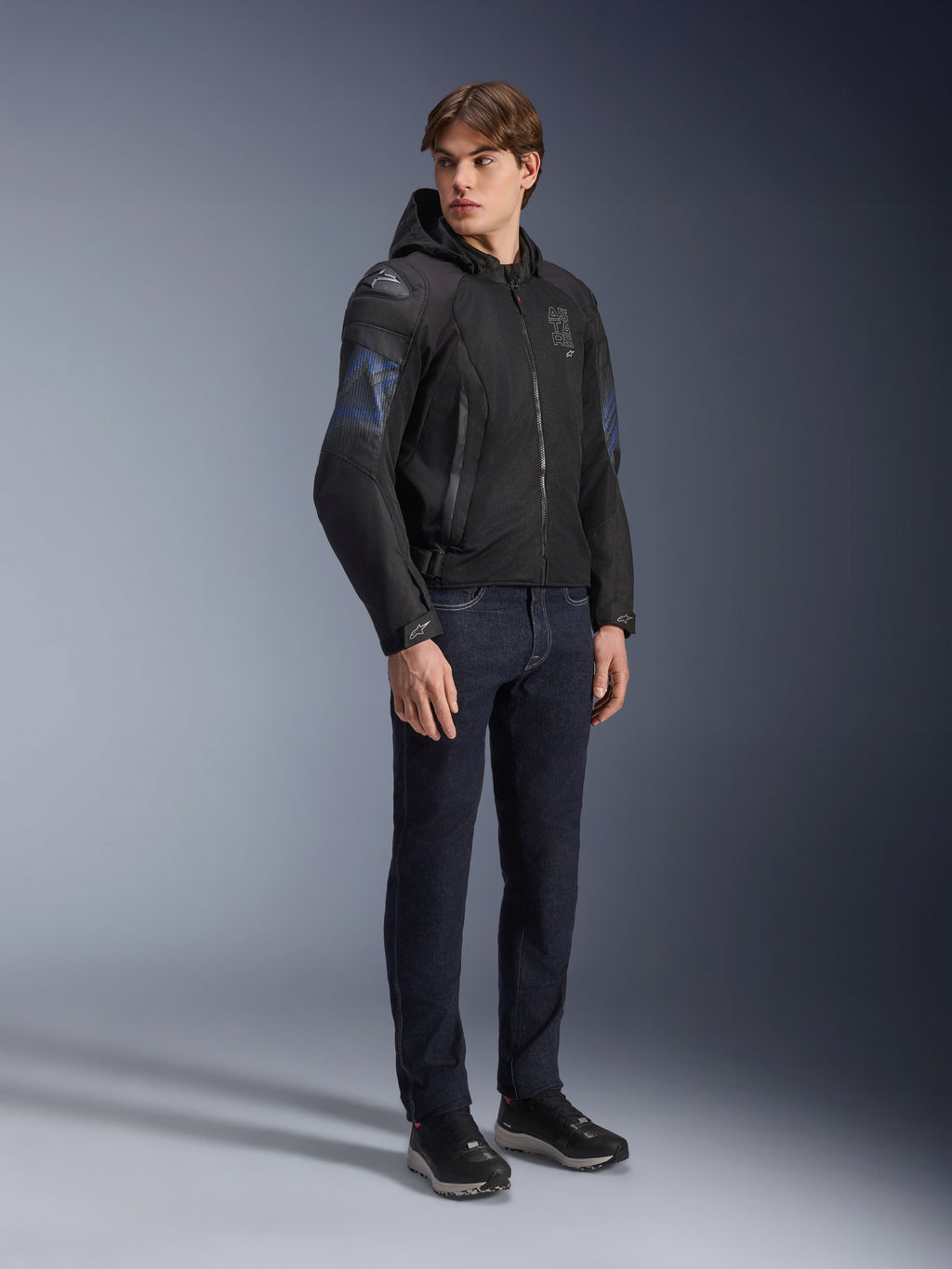Giacca Alpinestars Zaca Air Venom WP, giacca da moto in tessuto nero e blu elettrico, indossata da un modello con jeans scuri e sneakers nere, dotata di cappuccio rimovibile, pannelli di ventilazione in mesh e slider sulle spalle DFS Lite, abbigliamento tecnico sportivo per tutte le condizioni atmosferiche.