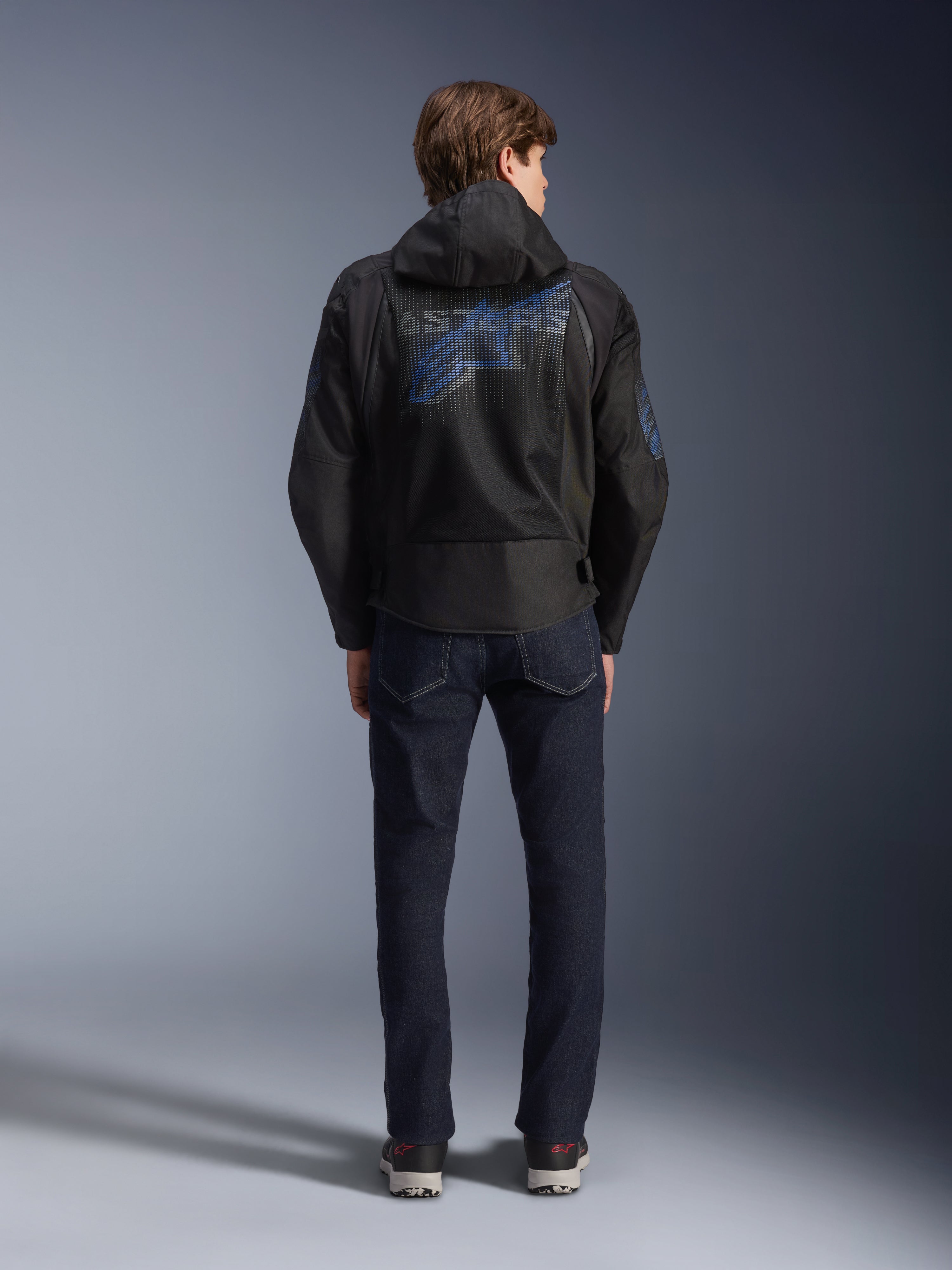 Giacca Alpinestars Zaca Air Venom WP, nero e blu elettrico, vista posteriore con un grande logo Alpinestars blu sul pannello posteriore in rete, cappuccio rimovibile, indossata con jeans in denim scuro e sneakers, abbigliamento da moto sportivo per tutte le stagioni.