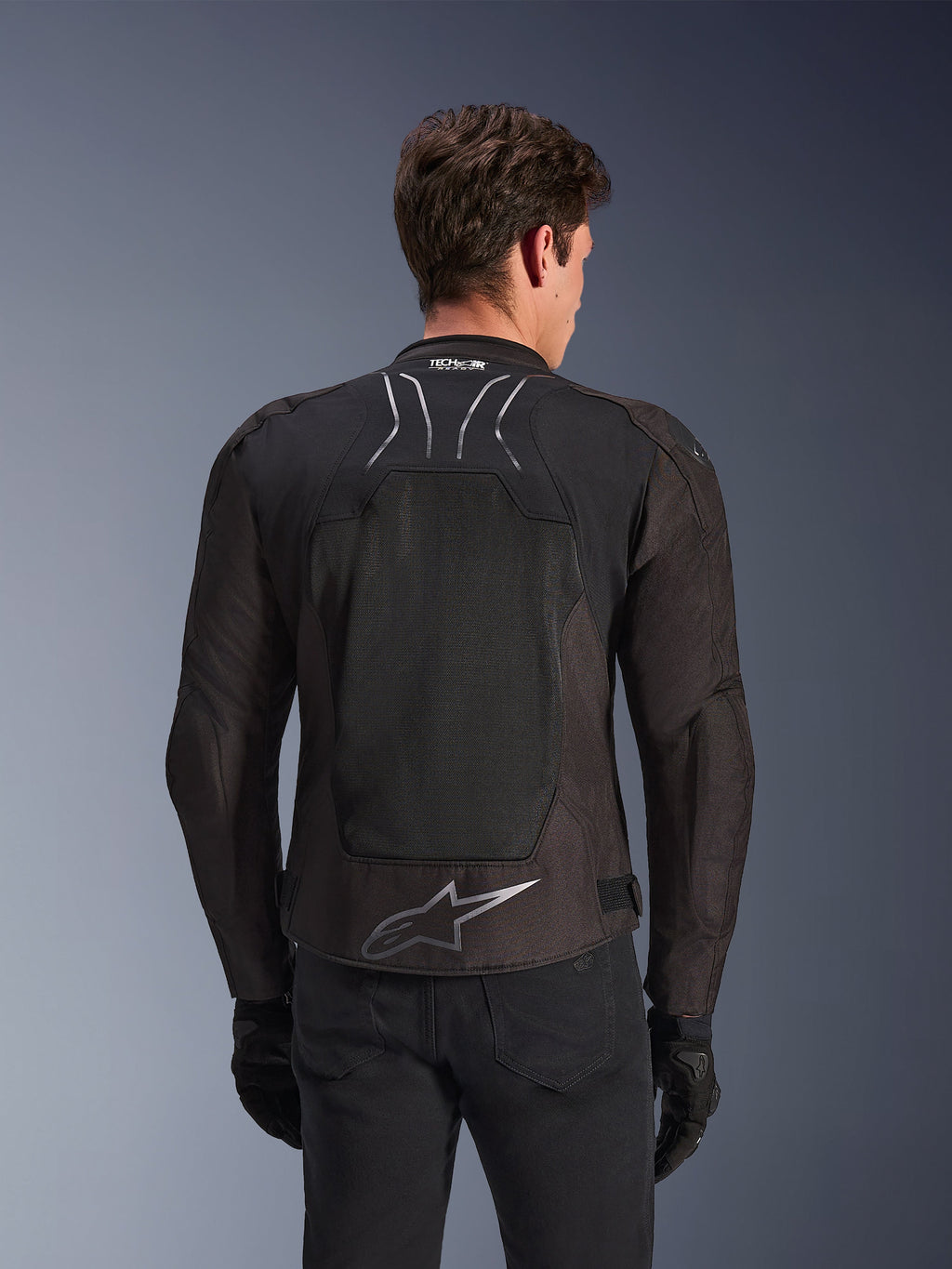 Giacca Alpinestars SMX Air in Black Anthracite, vista posteriore su un modello, ampio pannello posteriore in mesh tecnico per la ventilazione, marchio Tech-Air Ready sul colletto e logo Astar argento sulla parte bassa della schiena, abbinata a guanti neri e pantaloni da moto.