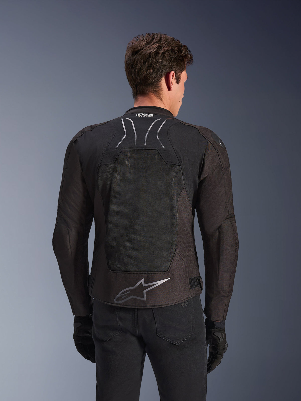 Giacca Alpinestars SMX Air in Black Anthracite, vista posteriore su un modello, ampio pannello posteriore in mesh tecnico per la ventilazione, marchio Tech-Air Ready sul colletto e logo Astar argento sulla parte bassa della schiena, abbinata a guanti neri e pantaloni da moto.