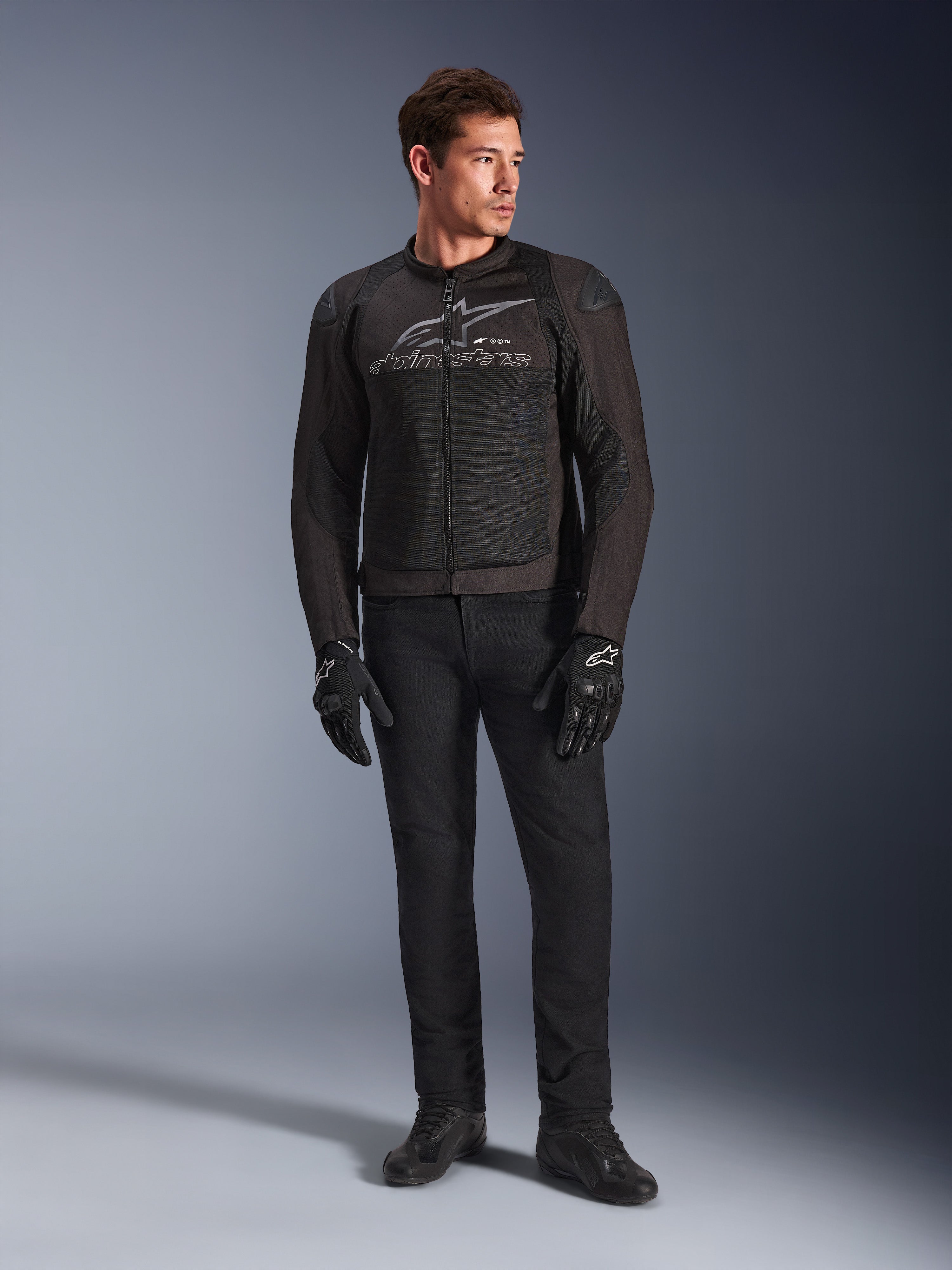 Modello indossa la giacca Alpinestars SMX Air, giacca da moto Black Anthracite con pannelli in rete ventilata e logo grigio sul petto, abbinata a pantaloni neri e guanti protettivi, ambientazione in studio con sfondo sfumato