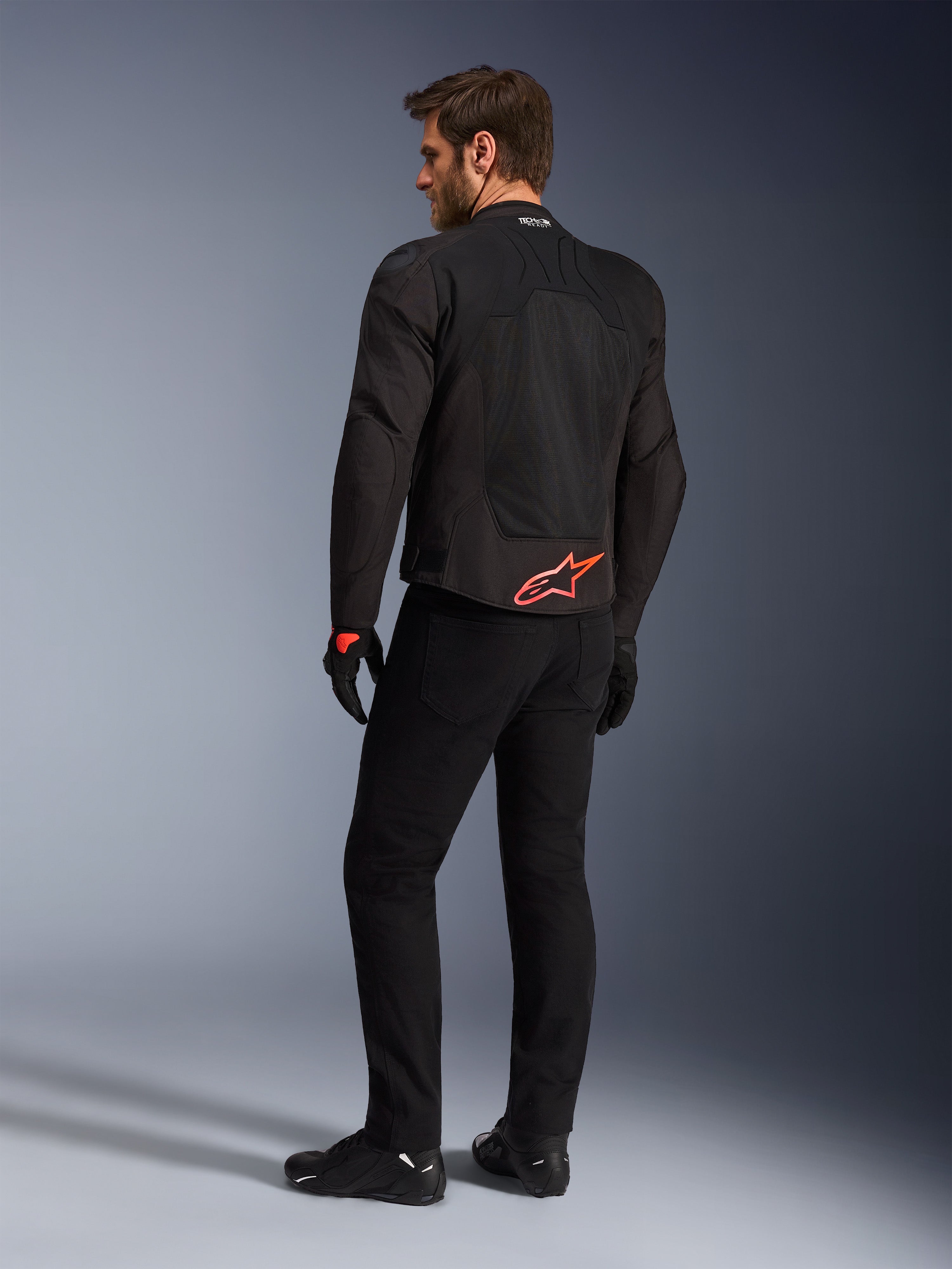 Vista posteriore di un modello che indossa la giacca Alpinestars SMX Air in nero, con logo Alpinestars rosso acceso sulla parte bassa della schiena, marchio 'Tech-Air Ready' sul colletto e pannelli in rete tecnica per la ventilazione, abbinata a pantaloni e guanti da moto neri.