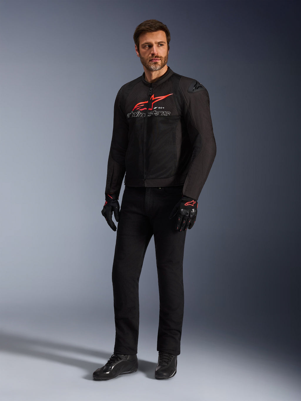 Modello uomo che indossa la giacca Alpinestars SMX Air in Nero Rosso Brillante Bianco, abbinata a pantaloni tecnici neri, guanti neri e rossi e scarpe da guida nere, con un evidente logo rosso sul petto e branding bianco, scatto in studio a figura intera per abbigliamento sportivo da motociclismo