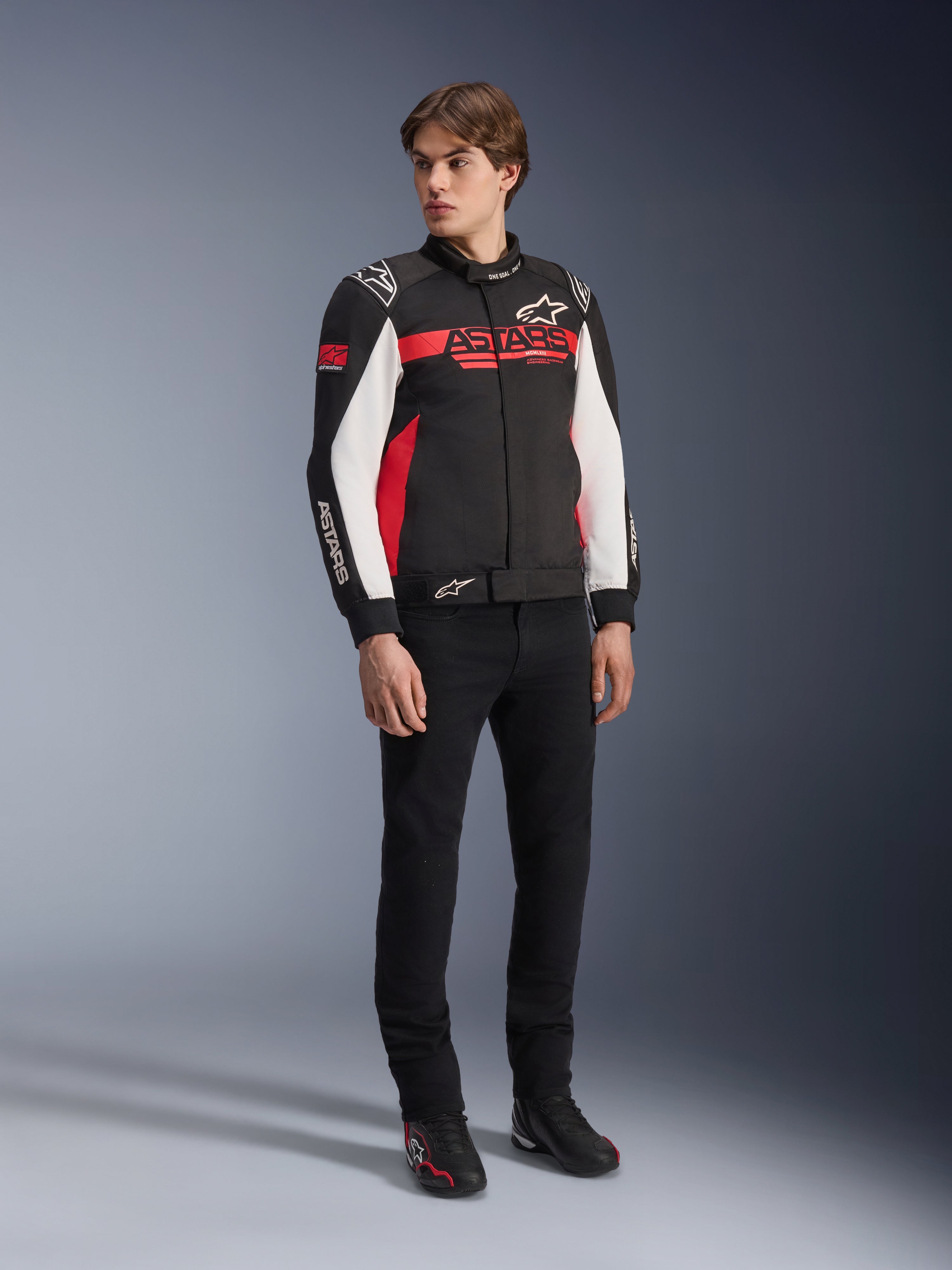 Giacca Alpinestars Monza Sport, nero, rosso acceso e bianco, giacca da moto in tessuto, design ispirato alle corse con marchio ASTARS su petto e maniche, indossata da un modello con pantaloni neri e scarpe da moto, abbigliamento da guida casual sportivo
