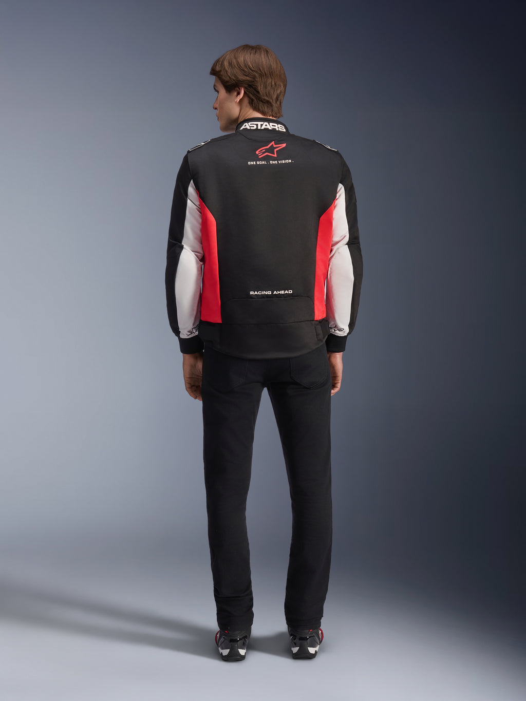 Giacca Alpinestars Monza Sport, giacca da moto in tessuto nero, rosso acceso e bianco, vista posteriore su un modello, con logo a stella rossa e marchio 'ASTARS' sul colletto con slogan, indossata con pantaloni neri e stivali da moto.