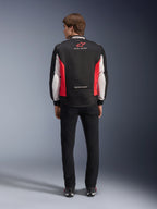 Giacca Alpinestars Monza Sport, giacca da moto in tessuto nero, rosso acceso e bianco, vista posteriore su un modello, con logo a stella rossa e marchio 'ASTARS' sul colletto con slogan, indossata con pantaloni neri e stivali da moto.