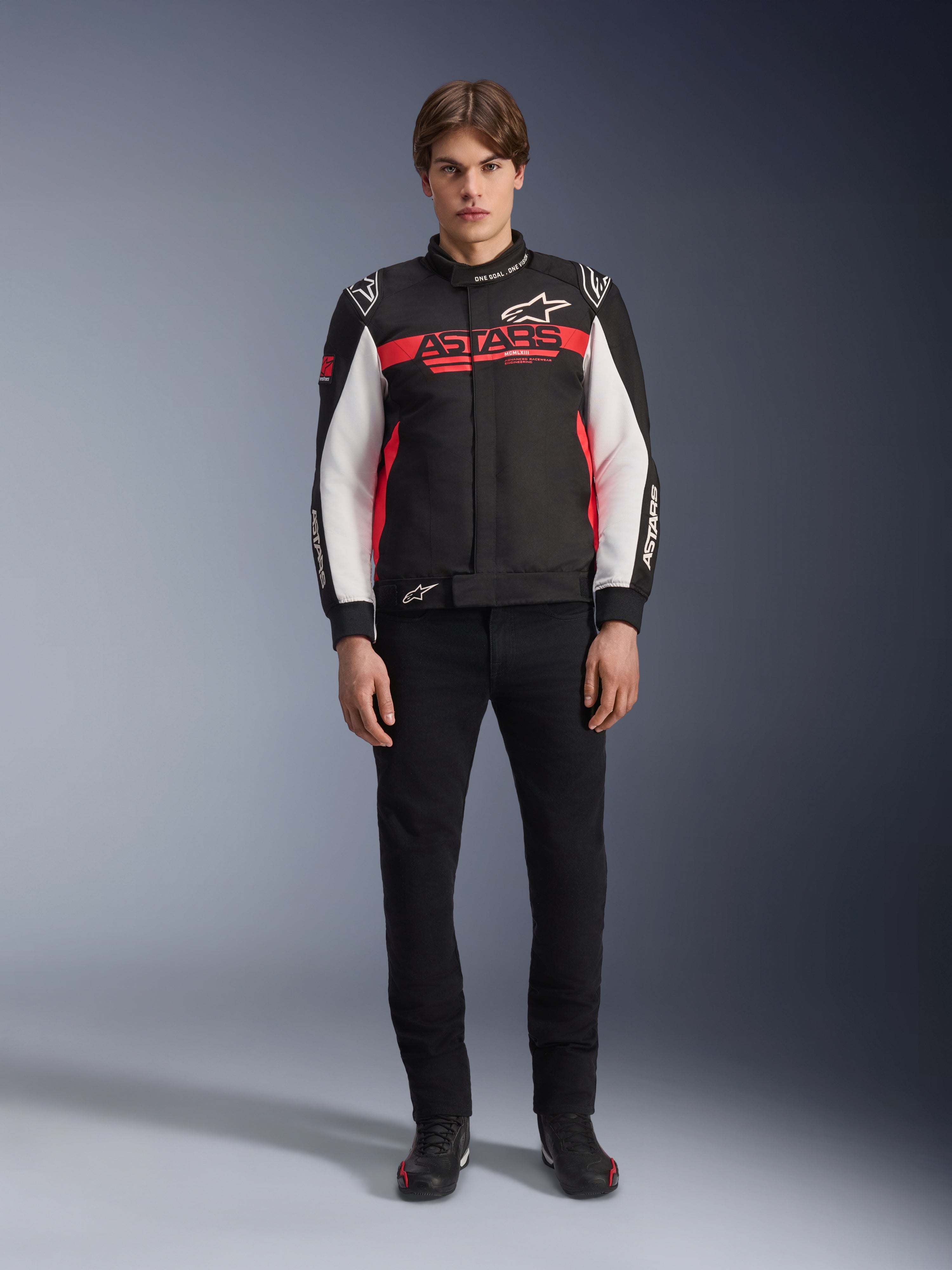 Modello che indossa la giacca Alpinestars Monza Sport in nero, rosso acceso e bianco, con un grande logo 'ASTARS' rosso sul petto e pannelli bianchi sulle maniche, abbinata a pantaloni neri e stivali da moto neri.