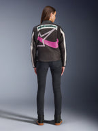 Vista posteriore di una donna che indossa la giacca Alpinestars Idles, nera con strisce bianco sporco, caratterizzata da un grande logo Alpinestars a stella viola e bianco e una scritta verde sul retro, abbinata a jeans slim-fit scuri, abbigliamento da moto urbano d'ispirazione vintage