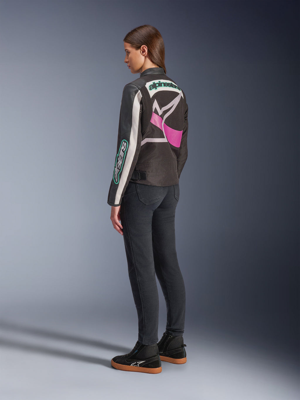 Giacca Alpinestars Idles da donna, giacca da moto, nero, bianco sporco e viola, vista dal retro su modella, con un grande logo Alpinestars avvolgente e design d'ispirazione racing vintage, abbinata a jeans scuri e scarpe nere.
