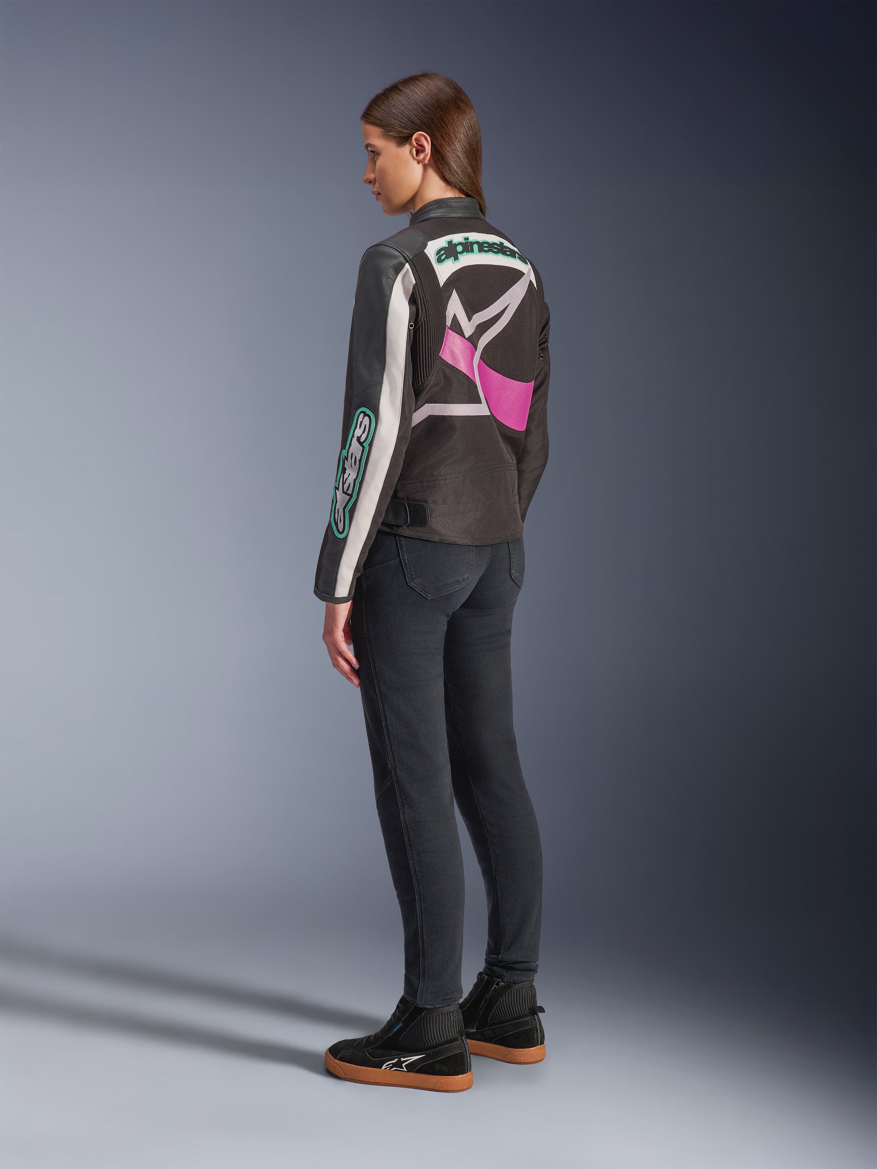 Giacca Alpinestars Idles da donna, giacca da moto, nero, bianco sporco e viola, vista dal retro su modella, con un grande logo Alpinestars avvolgente e design d'ispirazione racing vintage, abbinata a jeans scuri e scarpe nere.