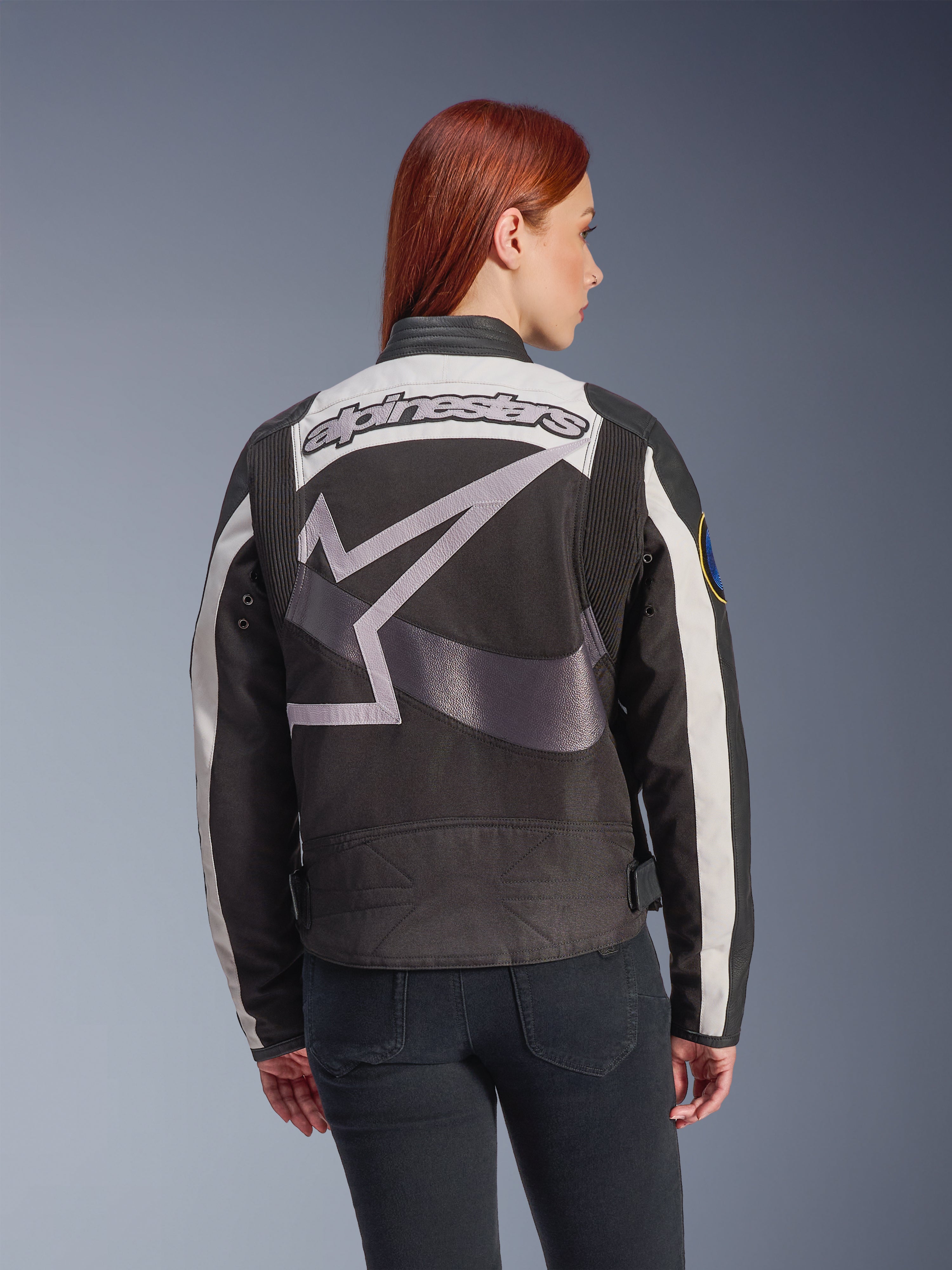 Giacca da donna Alpinestars Idles, nero e bianco sporco, vista posteriore con grande logo a stella grigio e scritta Alpinestars, indossata da una modella con i capelli rossi, giacca da moto urbana d'ispirazione vintage con pannelli a fisarmonica.