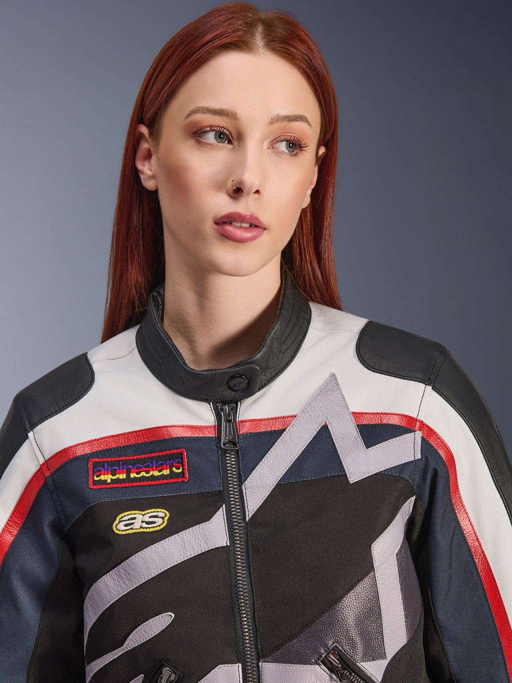 Giacca da donna Alpinestars Idles, primo piano di una modella con capelli rossi, colorazione Off White, Flame Red e Black, design racing d'ispirazione vintage con patch logo e grafiche a stella geometriche grigie, abbigliamento tecnico da moto urbano.