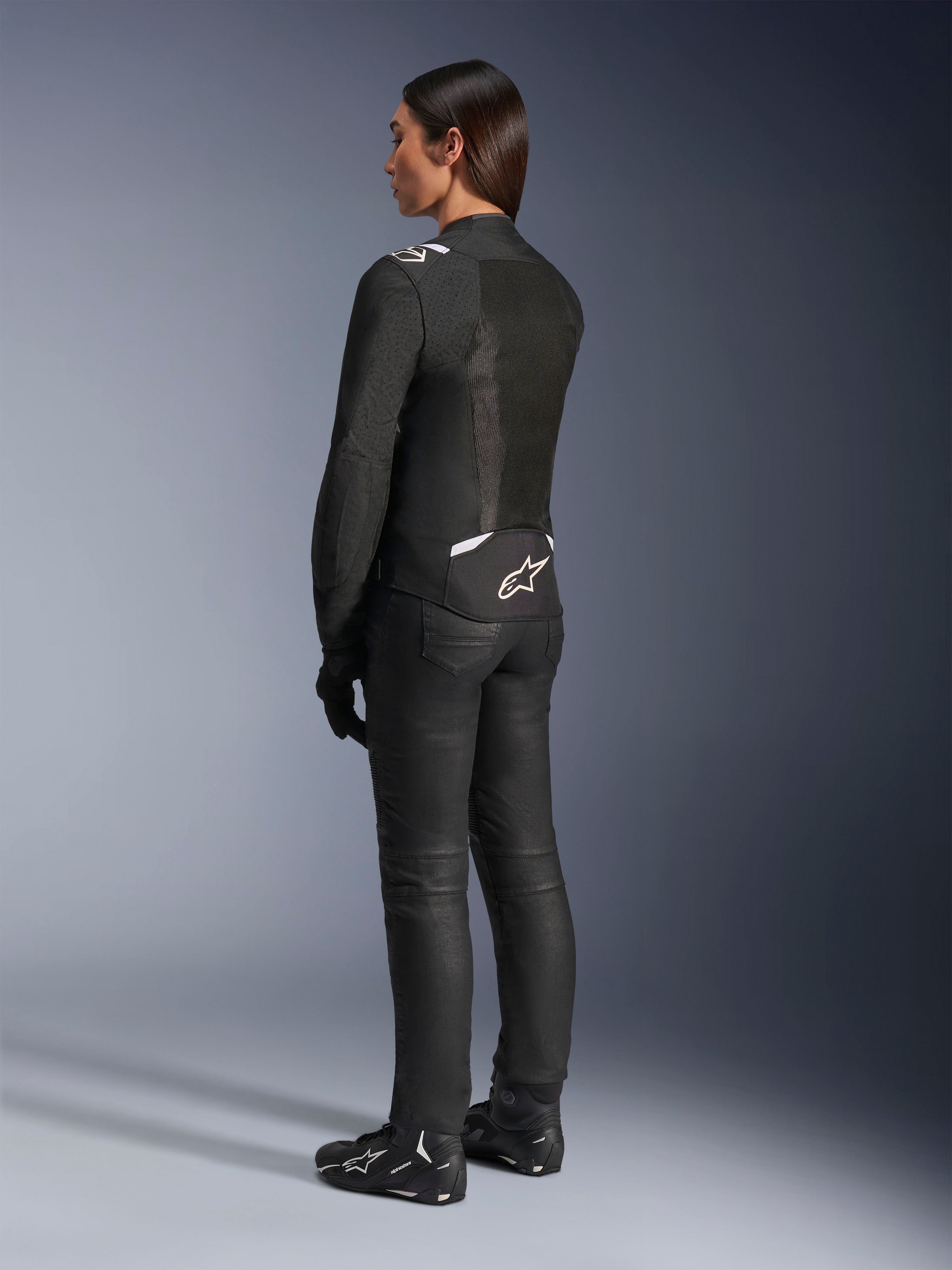 Giacca Alpinestars Stella T-Sps Air V2, nero e bianco, vista posteriore con pannelli in mesh e logo sulla parte bassa della schiena, abbinata a pantaloni da guida neri e scarpe da moto, abbigliamento tecnico sportivo da strada per donna