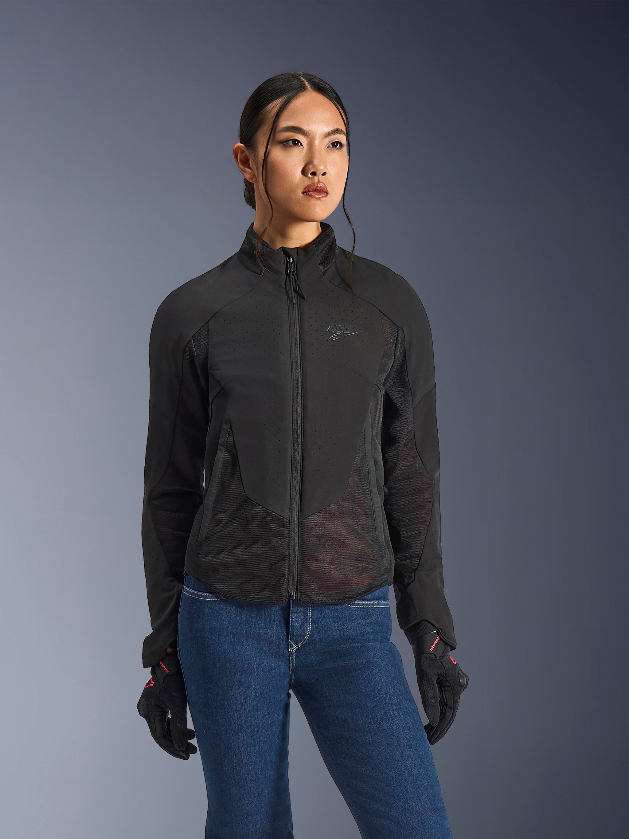 Giacca Alpinestars Stella Aeroshell Airflow, giacca da moto da donna, nero antracite, indossata da una modella con jeans blu e guanti neri, con softshell perforato al laser e pannelli in mesh per un'elevata ventilazione, design ergonomico per guida sportiva
