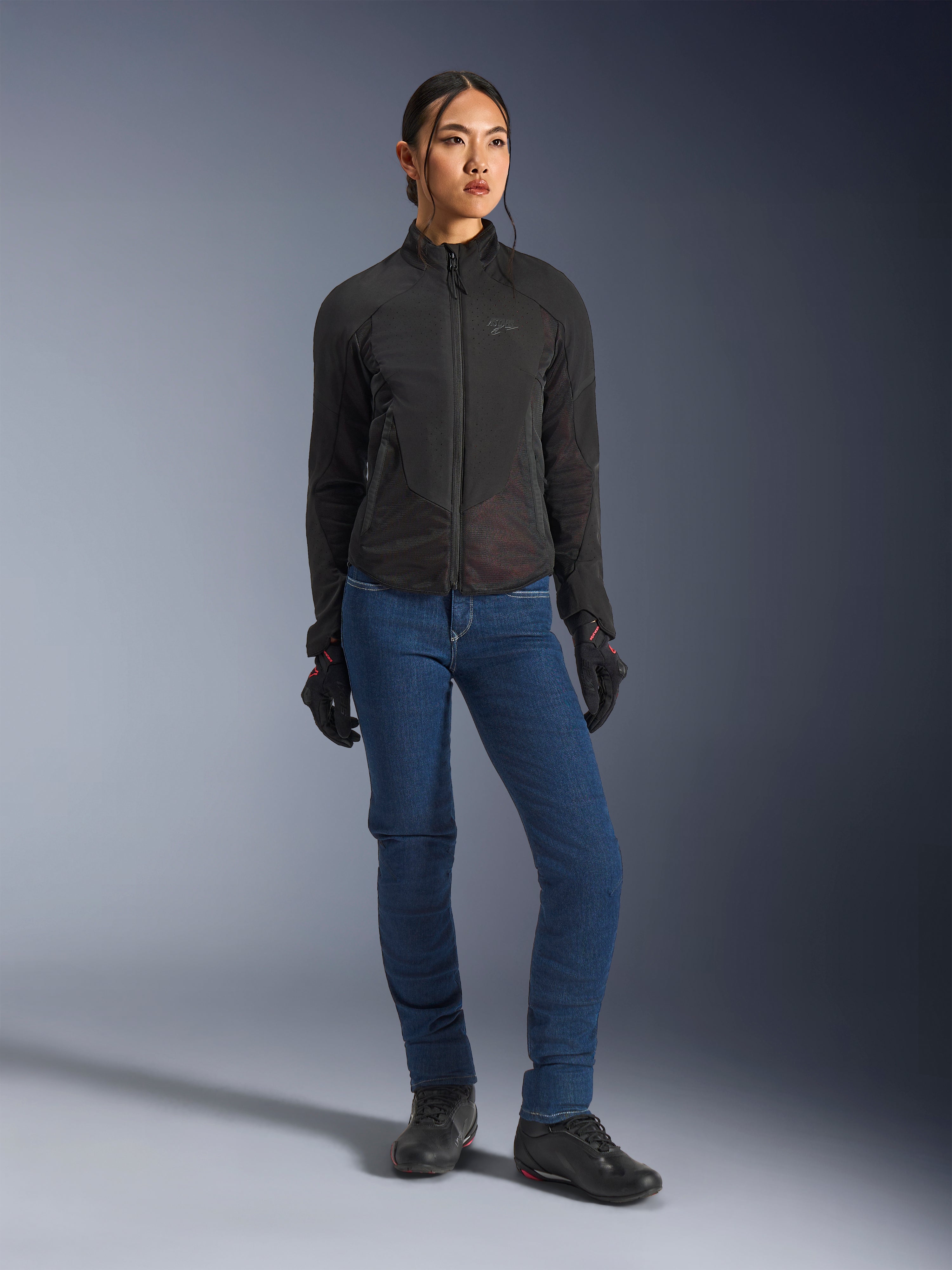 Motociclista con giacca Alpinestars Stella Aeroshell Airflow in nero, caratterizzata da ventilazione traforata al laser e pannelli in mesh, abbinata a jeans da moto blu e attrezzatura tecnica nera, estetica sportiva e urbana moderna