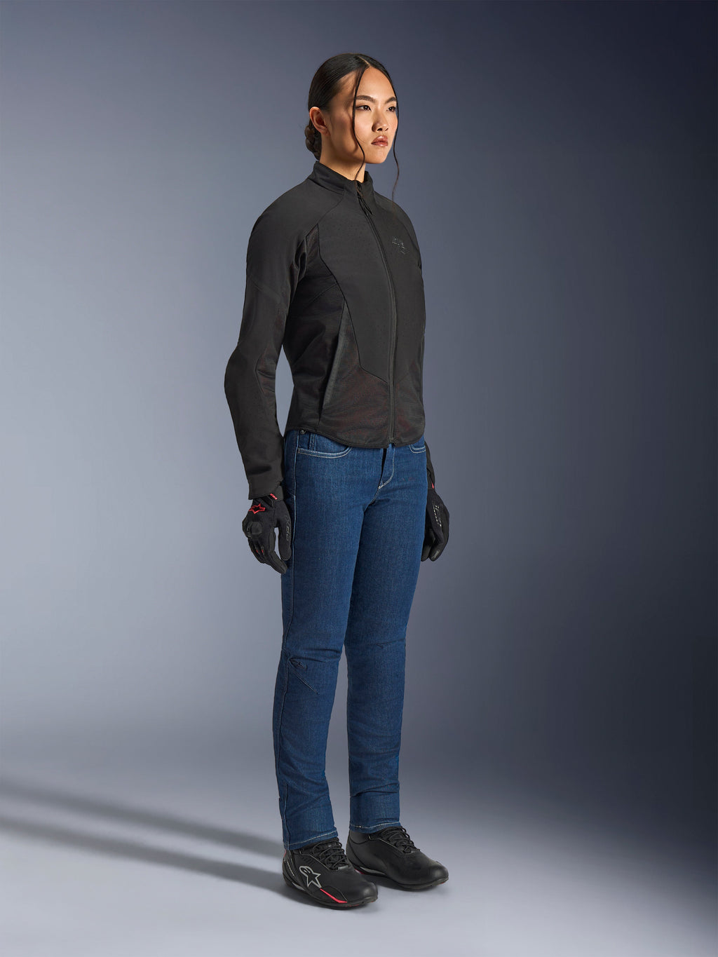 Motociclista che indossa la giacca Alpinestars Stella Aeroshell Airflow in Black Anthracite, giacca da moto softshell slim-fit, abbinata a jeans da guida blu e guanti neri, vista in studio a figura intera