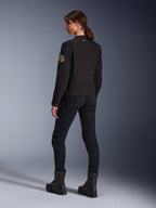 Modella che indossa la giacca Alpinestars Stella Grace in nero, vista posteriore che mette in risalto la toppa vintage Oscar a spirale sul braccio e il piccolo logo a stella sul colletto, abbinata a pantaloni da moto e stivali neri per la guida urbana