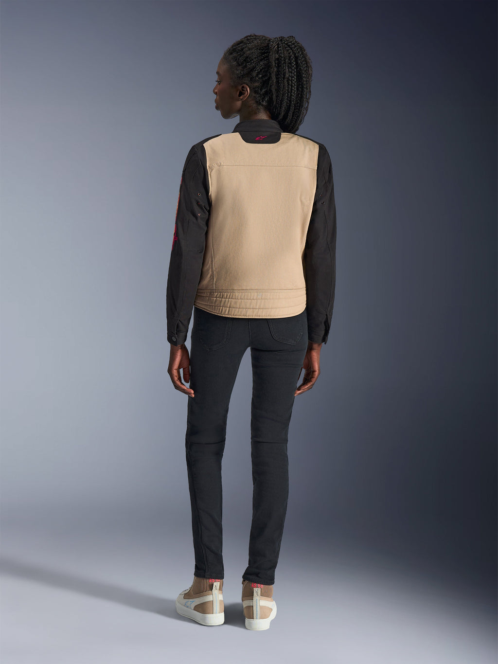 Donna che indossa la giacca Alpinestars Stella Grace, Aluminum Black con busto tan e maniche nere, vista posteriore, abbinata a pantaloni neri slim-fit e sneakers casual, abbigliamento da moto urbano da donna