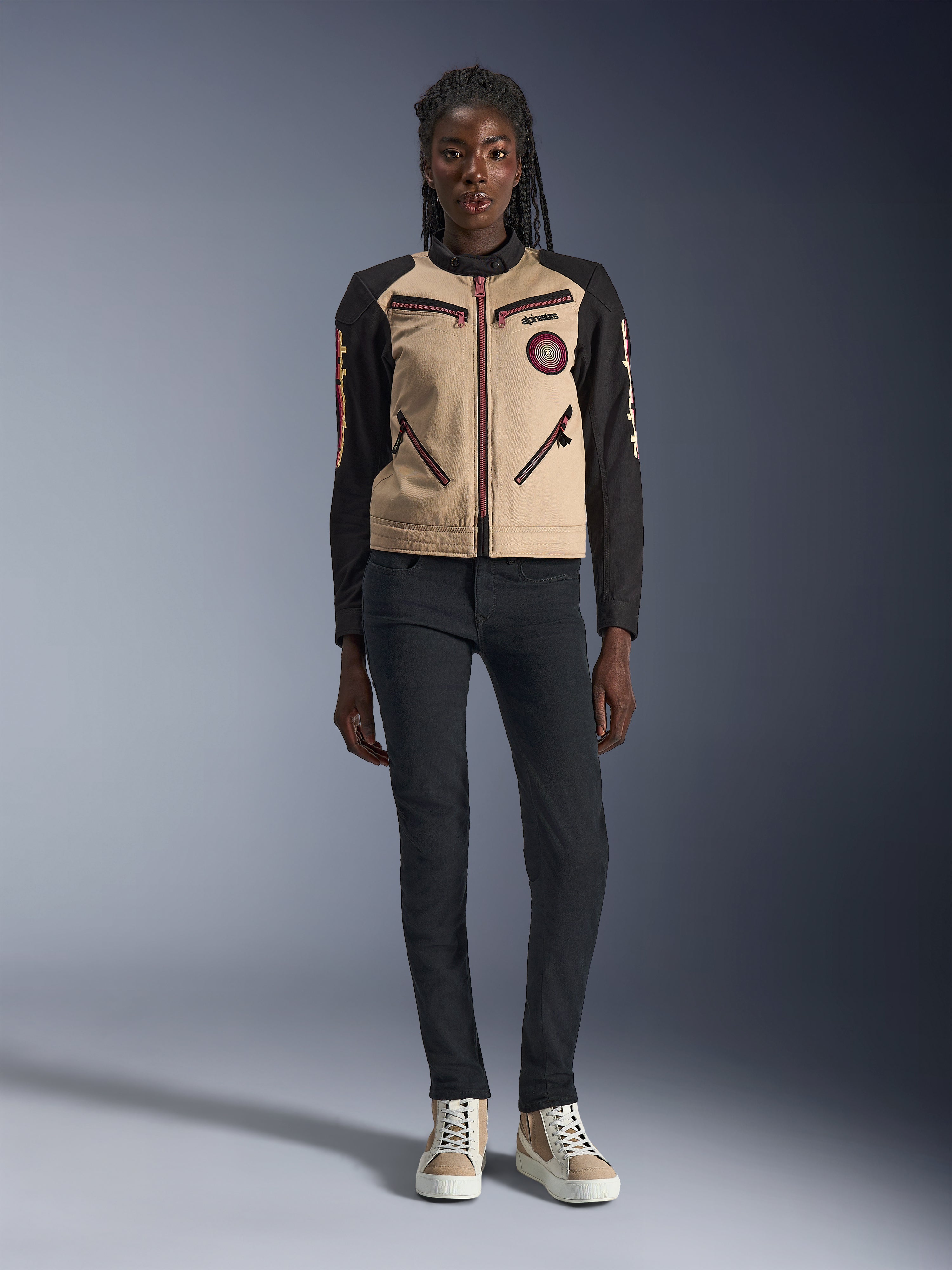 Giacca Alpinestars Stella Grace da donna, giacca da moto urban, costruzione in tessuto color sabbia e nero, con cerniere anteriori e patch Oscar a spirale ricamata, indossata con pantaloni neri e sneaker alte, abbigliamento tecnico da moto ispirato al workwear contemporaneo