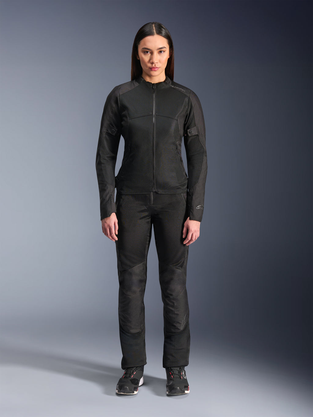 Donna che indossa la giacca Alpinestars Stella C-1 Air in nero, abbinata a pantaloni da moto in tessuto nero coordinati e scarpe da guida, caratterizzata da un design urbano elegante con ampi pannelli in mesh per il flusso d'aria.
