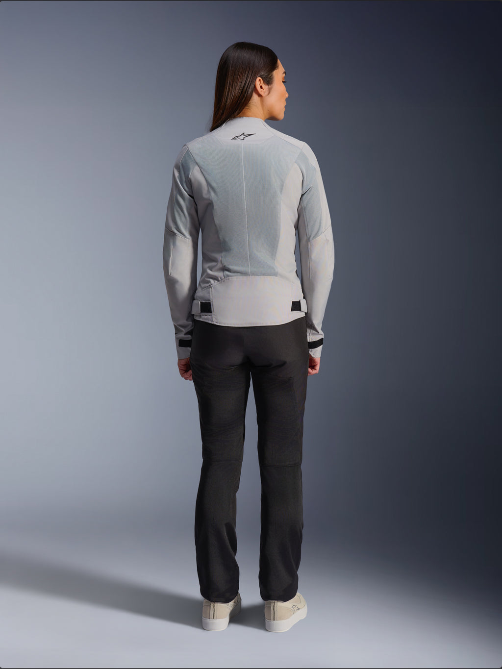 Donna che indossa la giacca Alpinestars Stella C-1 Air in Silver, vista posteriore, caratterizzata da ampi pannelli in rete tecnica per la ventilazione e un logo a stella riflettente, abbinata a pantaloni da moto neri e sneakers bianche, abbigliamento da moto urban contemporary