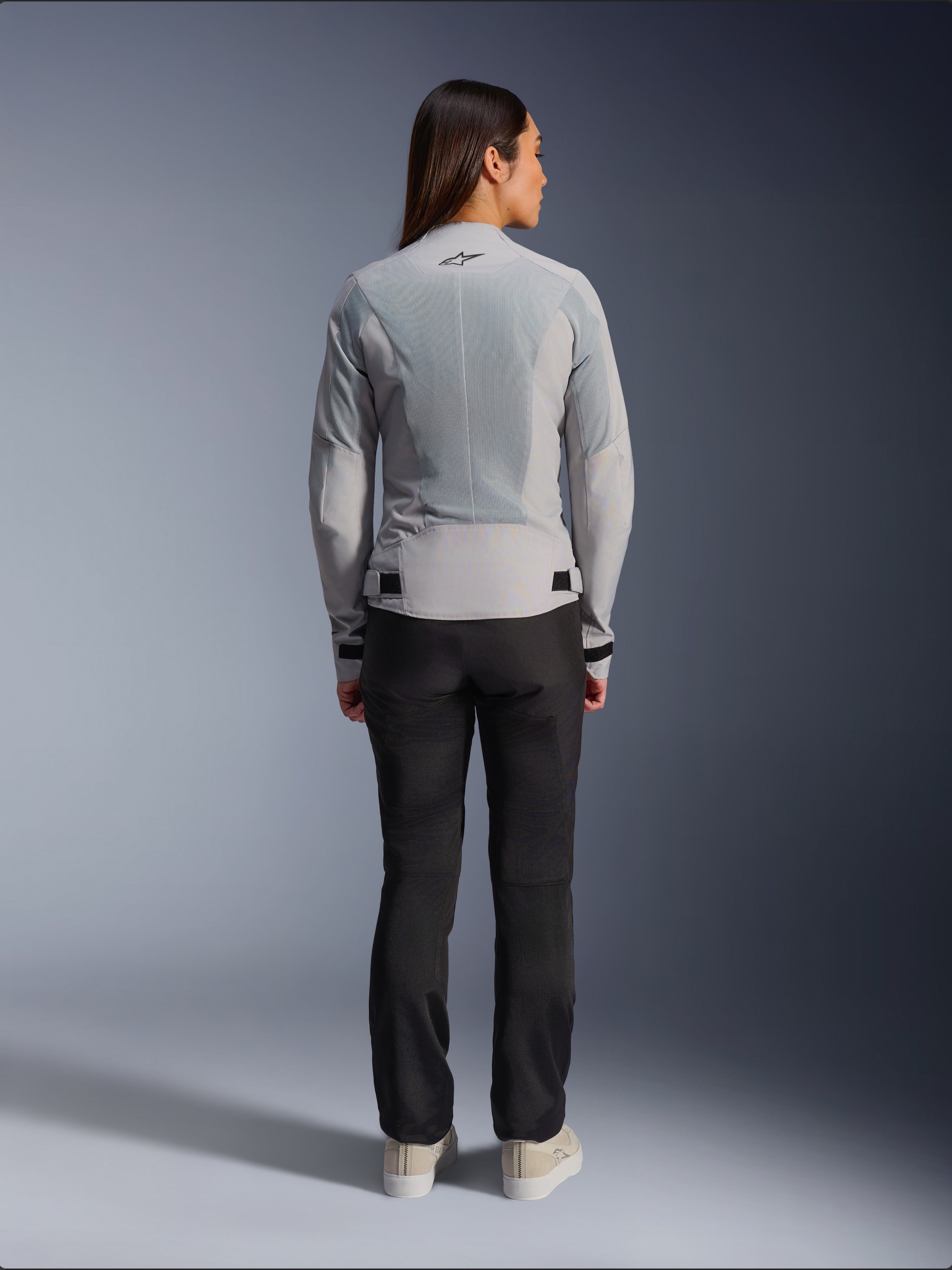 Donna che indossa la giacca Alpinestars Stella C-1 Air in Silver, vista posteriore, caratterizzata da ampi pannelli in rete tecnica per la ventilazione e un logo a stella riflettente, abbinata a pantaloni da moto neri e sneakers bianche, abbigliamento da moto urban contemporary