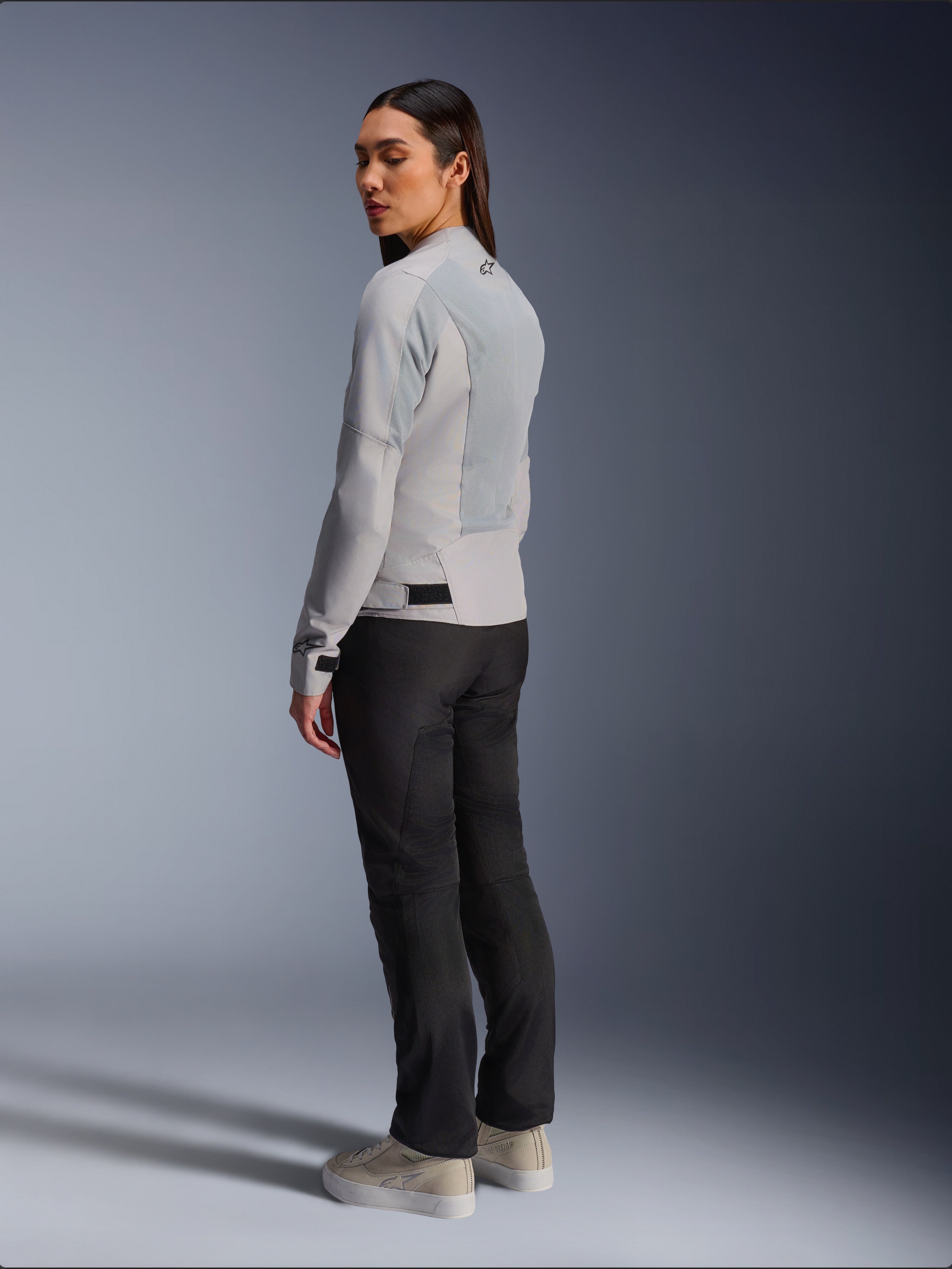 Donna che indossa la giacca Alpinestars Stella C-1 Air in Silver, vista posteriore e laterale che mostra i pannelli in rete tecnica e il logo riflettente sulla spalla, abbinata a pantaloni da moto neri e sneaker beige, estetica urban riding contemporanea