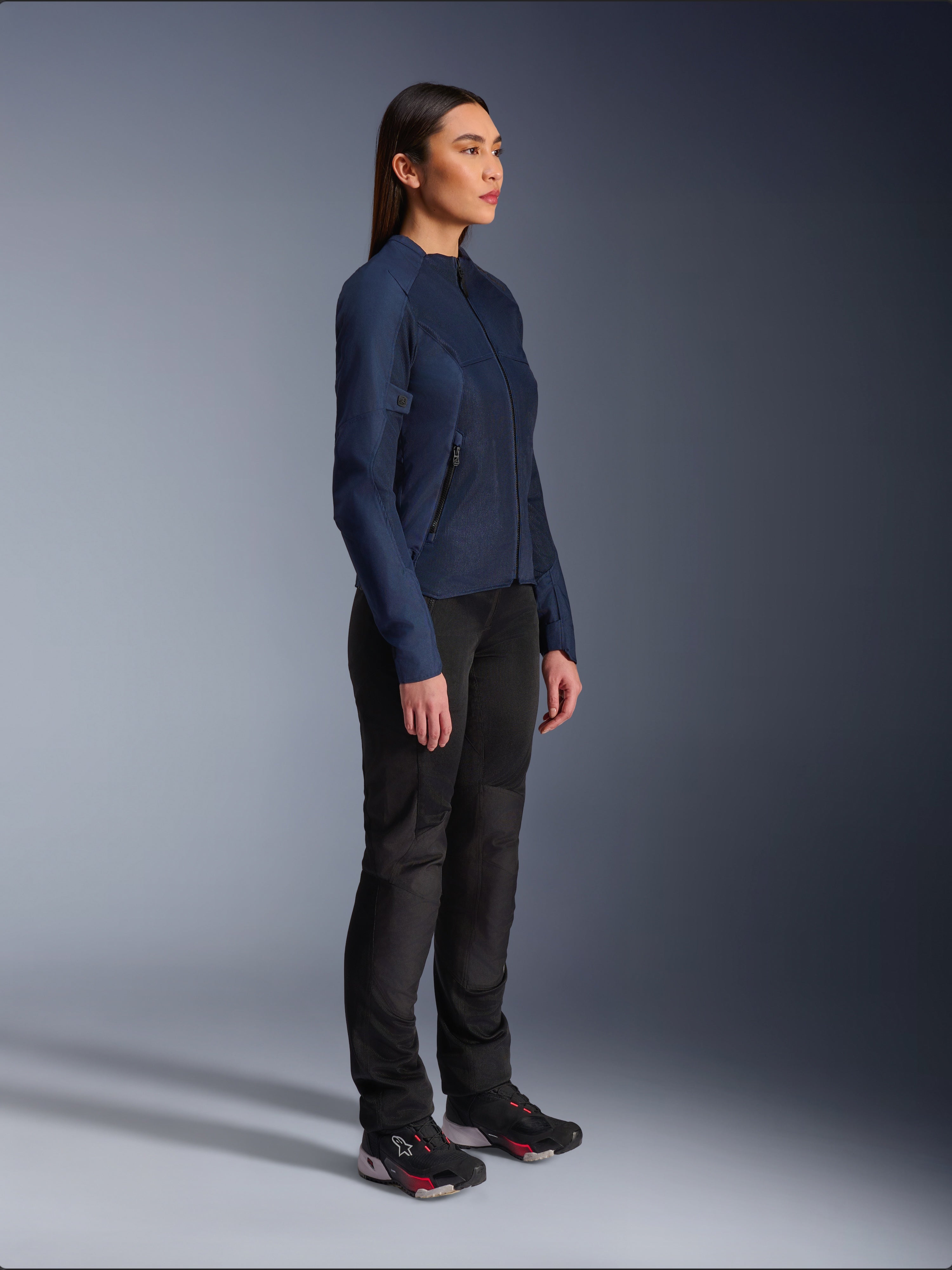 Donna che indossa la giacca Alpinestars Stella C-1 Air in blu, giacca da moto urban con pannelli in rete leggera per la ventilazione, abbinata a pantaloni da moto neri e scarpe tecniche, caratterizzata da un profilo snello e branding raffinato, vista laterale in studio