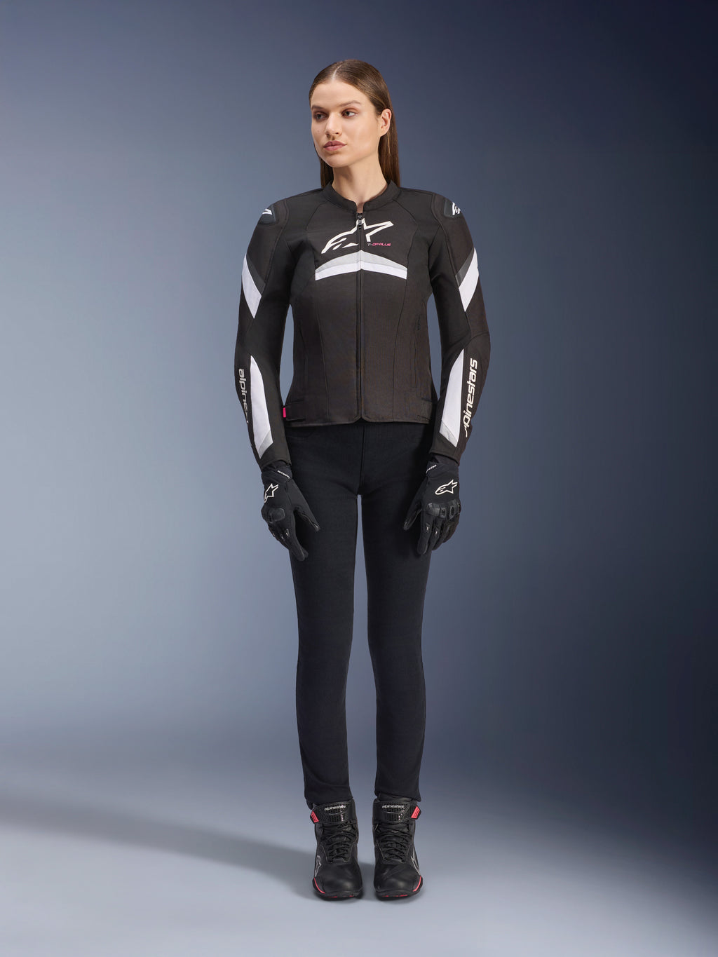 Modella che indossa la giacca Alpinestars Stella T-GP Plus R V4 in nero e bianco, giacca da moto sportiva in tessuto, abbinata a pantaloni da moto, guanti e stivali neri, vista frontale con logo Alpinestars bianco e dettagli sulle maniche.