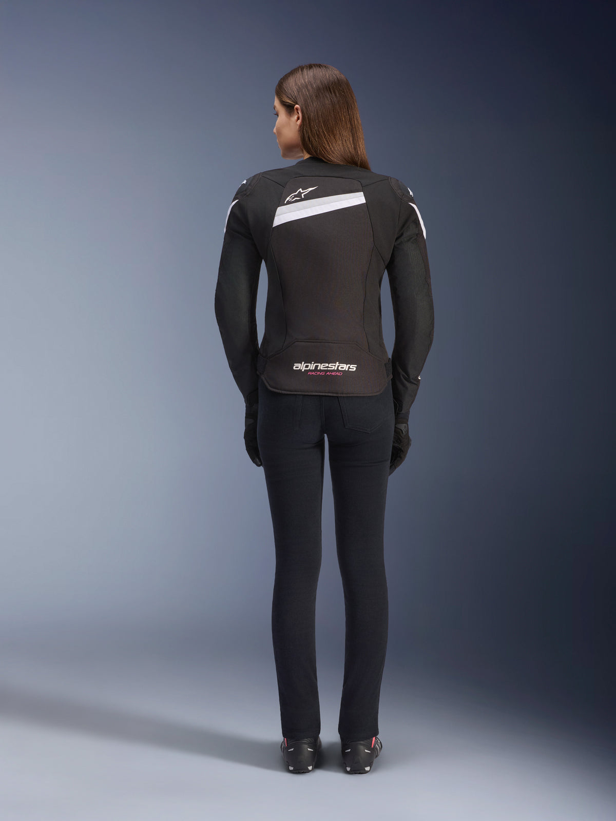 Giacca Alpinestars Stella T-GP Plus R V4, giacca da moto in tessuto da donna, nero e bianco, vista posteriore con accenti bianchi e loghi sulla schiena, indossata con pantaloni neri, vestibilità sportiva per prestazioni su strada