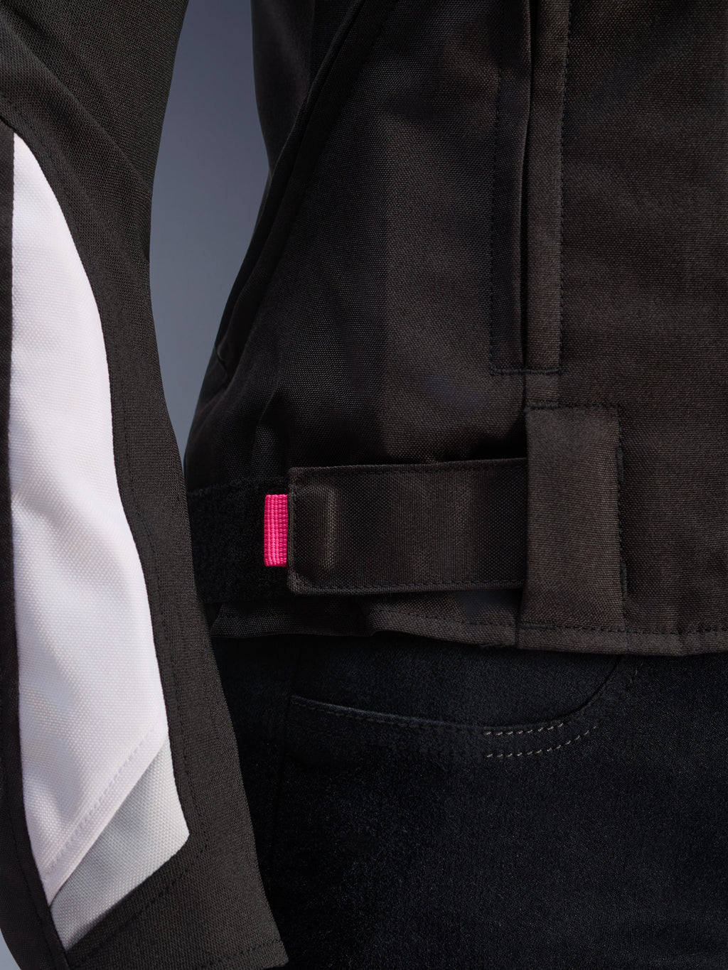 Giacca Alpinestars Stella T-GP Plus R V4, nero e bianco, primo piano del cinturino di regolazione in vita con linguetta rosa, robusta costruzione in tessuto con pannelli bianchi sulle maniche, giacca da moto da donna con vestibilità sportiva