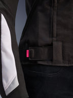 Giacca Alpinestars Stella T-GP Plus R V4, nero e bianco, primo piano del cinturino di regolazione in vita con linguetta rosa, robusta costruzione in tessuto con pannelli bianchi sulle maniche, giacca da moto da donna con vestibilità sportiva