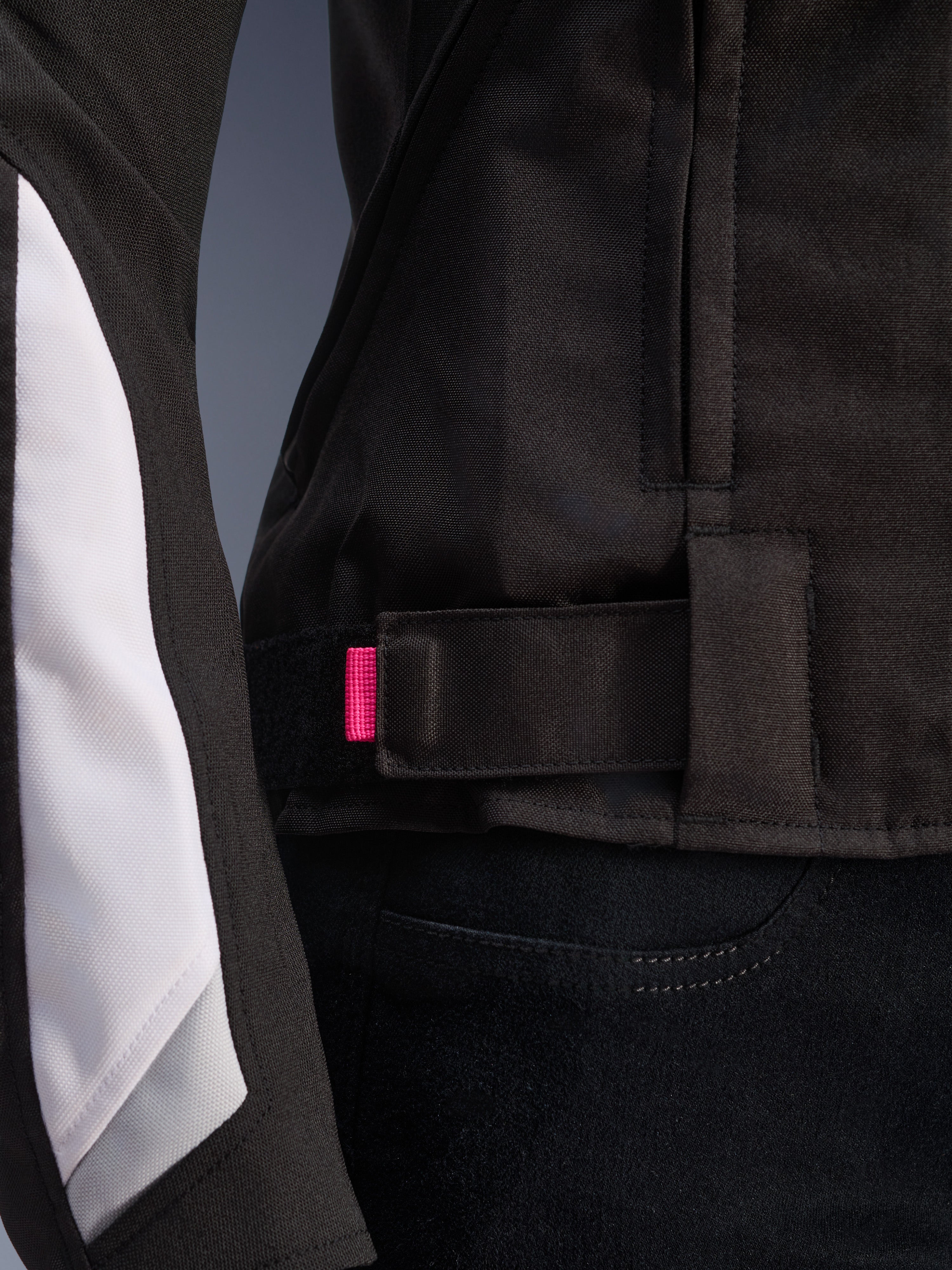 Giacca Alpinestars Stella T-GP Plus R V4, nero e bianco, primo piano del cinturino di regolazione in vita con linguetta rosa, robusta costruzione in tessuto con pannelli bianchi sulle maniche, giacca da moto da donna con vestibilità sportiva