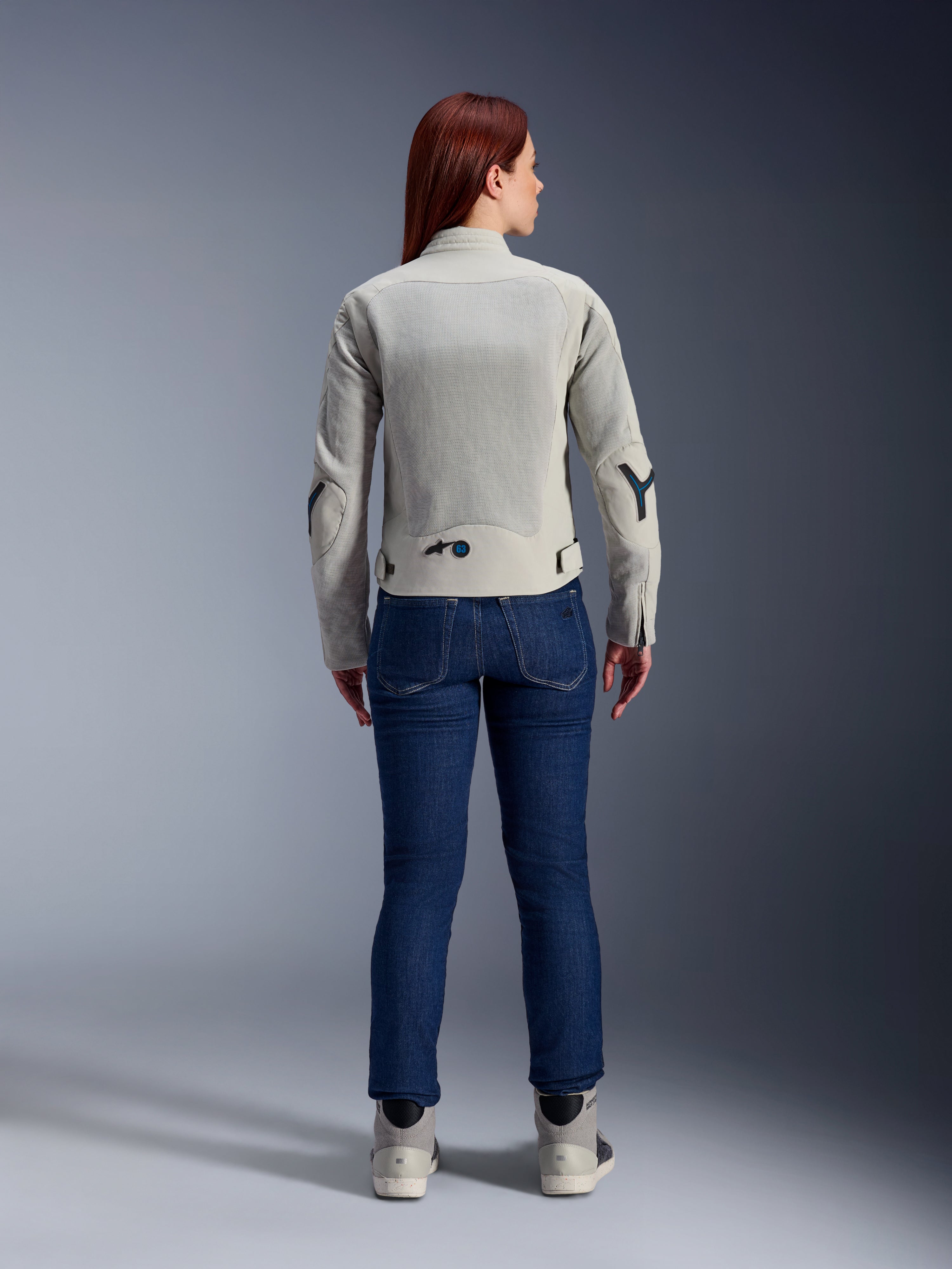 Giacca Alpinestars Stella Circal-1 Air da donna in grigio metallo e blu metile, vista posteriore, con ampi pannelli in rete per il flusso d'aria su schiena e braccia, indossata con jeans in denim blu scuro e scarpe da moto grigie per un look urban moderno.