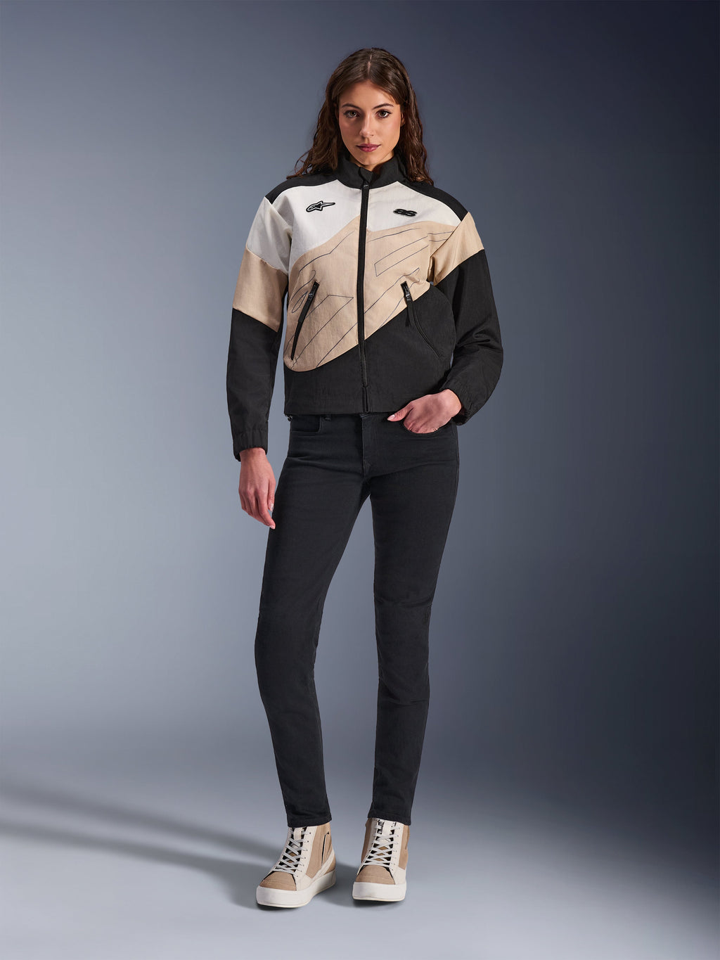 Giacca Alpinestars Stella Moshell da donna, giacca da moto urbana con design asimmetrico color-block in nero, sabbia e bianco sporco, indossata con pantaloni slim-fit neri e sneakers alte color sabbia, estetica streetwear moderna per motocicliste.