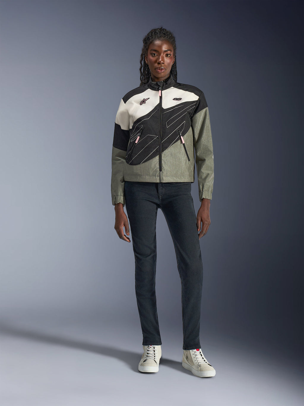 Giacca Alpinestars Stella Moshell, giacca da moto da donna, nero, grigio e bianco sporco con accenti rosa, indossata da una modella con jeans scuri e sneakers bianco sporco, caratterizzata da un design con zip asimmetrica e grafiche geometriche audaci, abbigliamento urbano contemporaneo