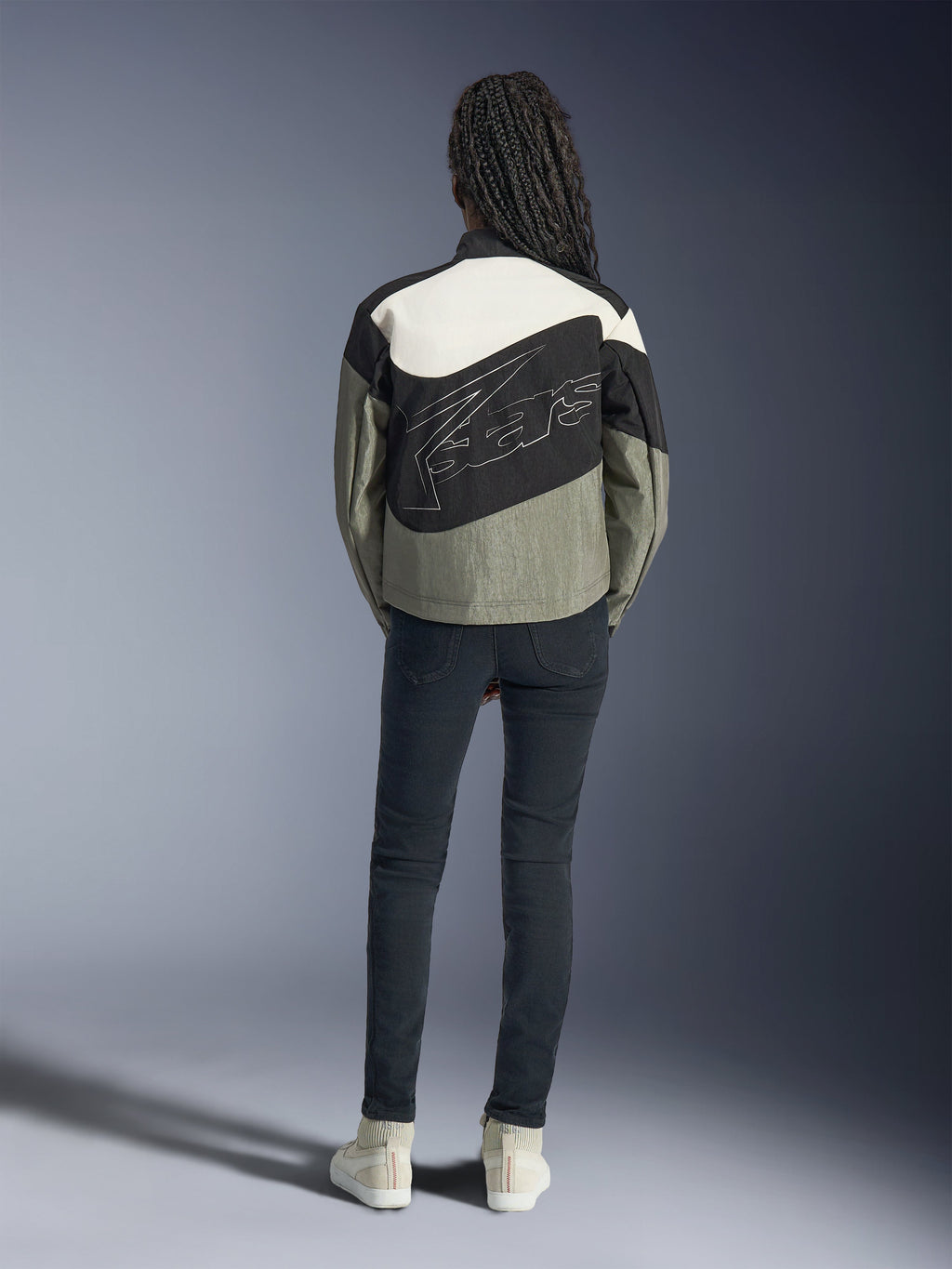 Vista posteriore di una modella che indossa la giacca Alpinestars Stella Moshell in nero, grigio e bianco sporco, caratterizzata da un design asimmetrico e un grande logo stilizzato profilato sul retro, abbinata a jeans scuri slim-fit e sneakers bianche, abbigliamento da moto per donna ispirato allo streetwear urbano.