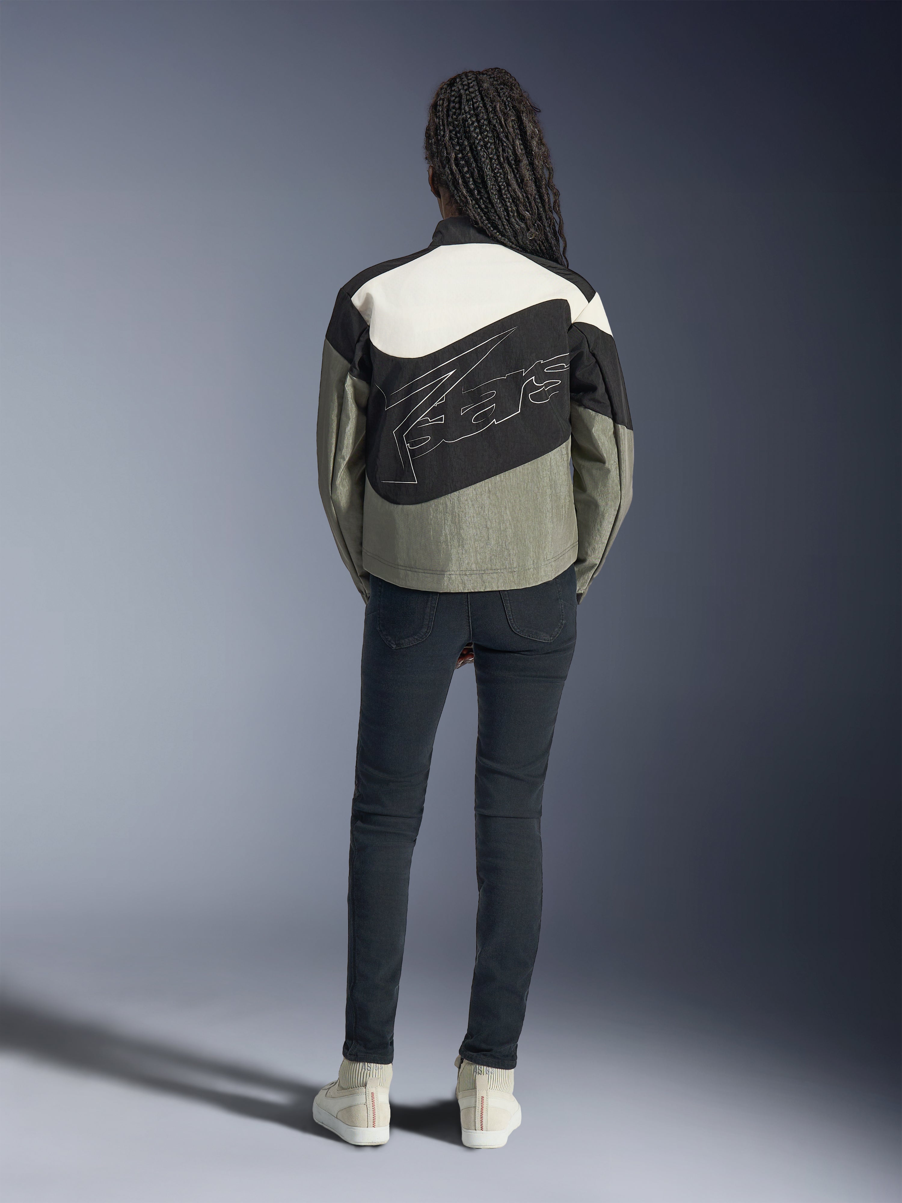 Vista posteriore di una modella che indossa la giacca Alpinestars Stella Moshell in nero, grigio e bianco sporco, caratterizzata da un design asimmetrico e un grande logo stilizzato profilato sul retro, abbinata a jeans scuri slim-fit e sneakers bianche, abbigliamento da moto per donna ispirato allo streetwear urbano.