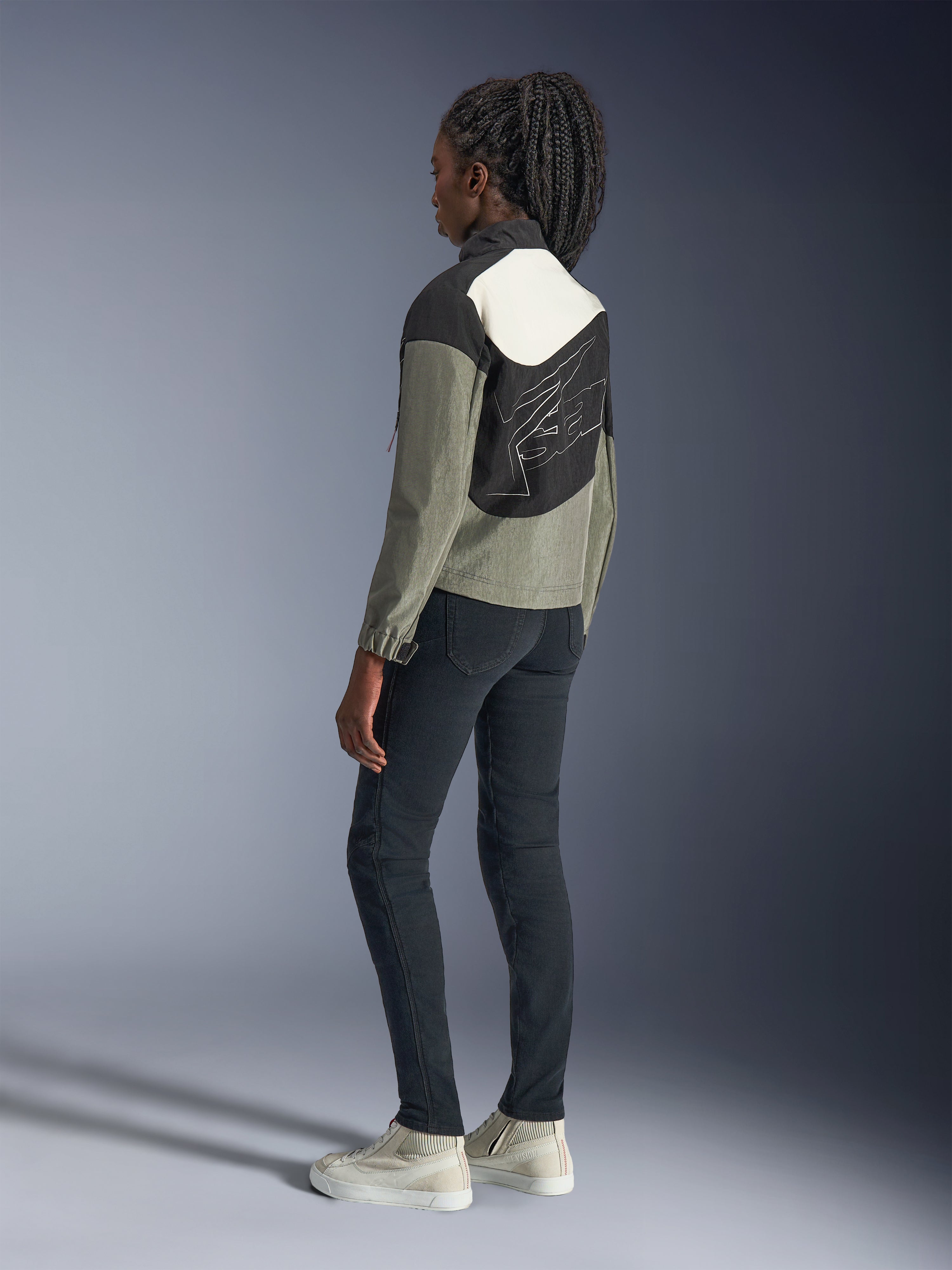 Donna che indossa la giacca Alpinestars Stella Moshell, giacca da moto urban, colorazione nero, grigio e bianco sporco, vista posteriore con design a pannelli asimmetrici e grande grafica sul retro, abbinata a pantaloni neri slim-fit e sneakers bianco sporco, abbigliamento da moto urban contemporaneo