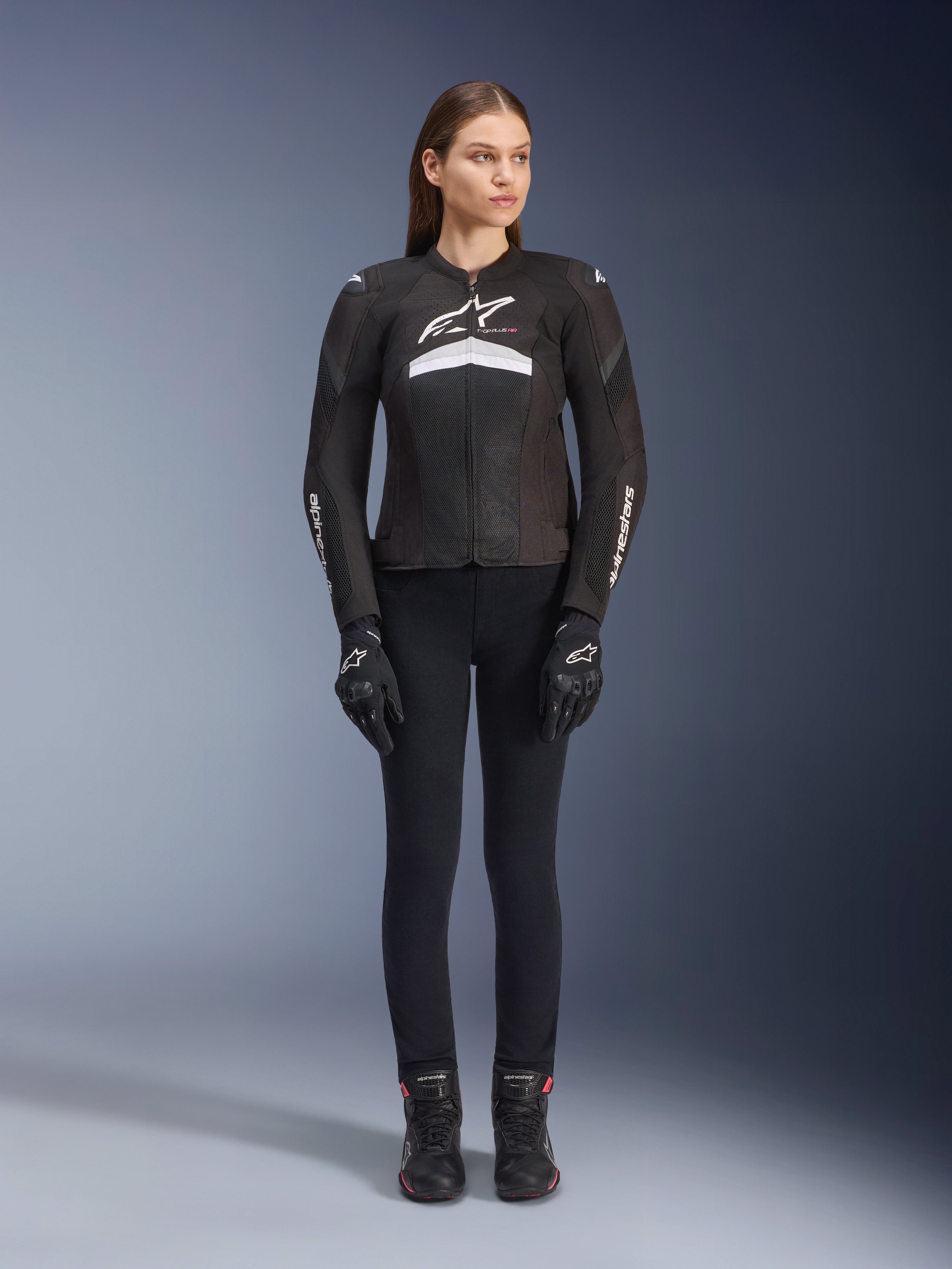 Motociclista donna che indossa la giacca Stella T-GP Plus R V4 Airflow, nero e bianco, abbinata a pantaloni da moto, guanti e stivali neri, con pannelli in mesh per la ventilazione e loghi Alpinestars bianchi su petto e braccia