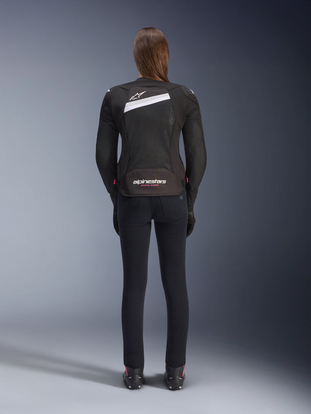 Giacca Alpinestars Stella T-GP Plus R V4 Airflow, nero e bianco, vista posteriore con ampi pannelli di ventilazione in mesh e marchio Alpinestars, indossata da una pilota con pantaloni da moto neri slim-fit e stivali, abbigliamento tecnico sportivo in tessuto da donna