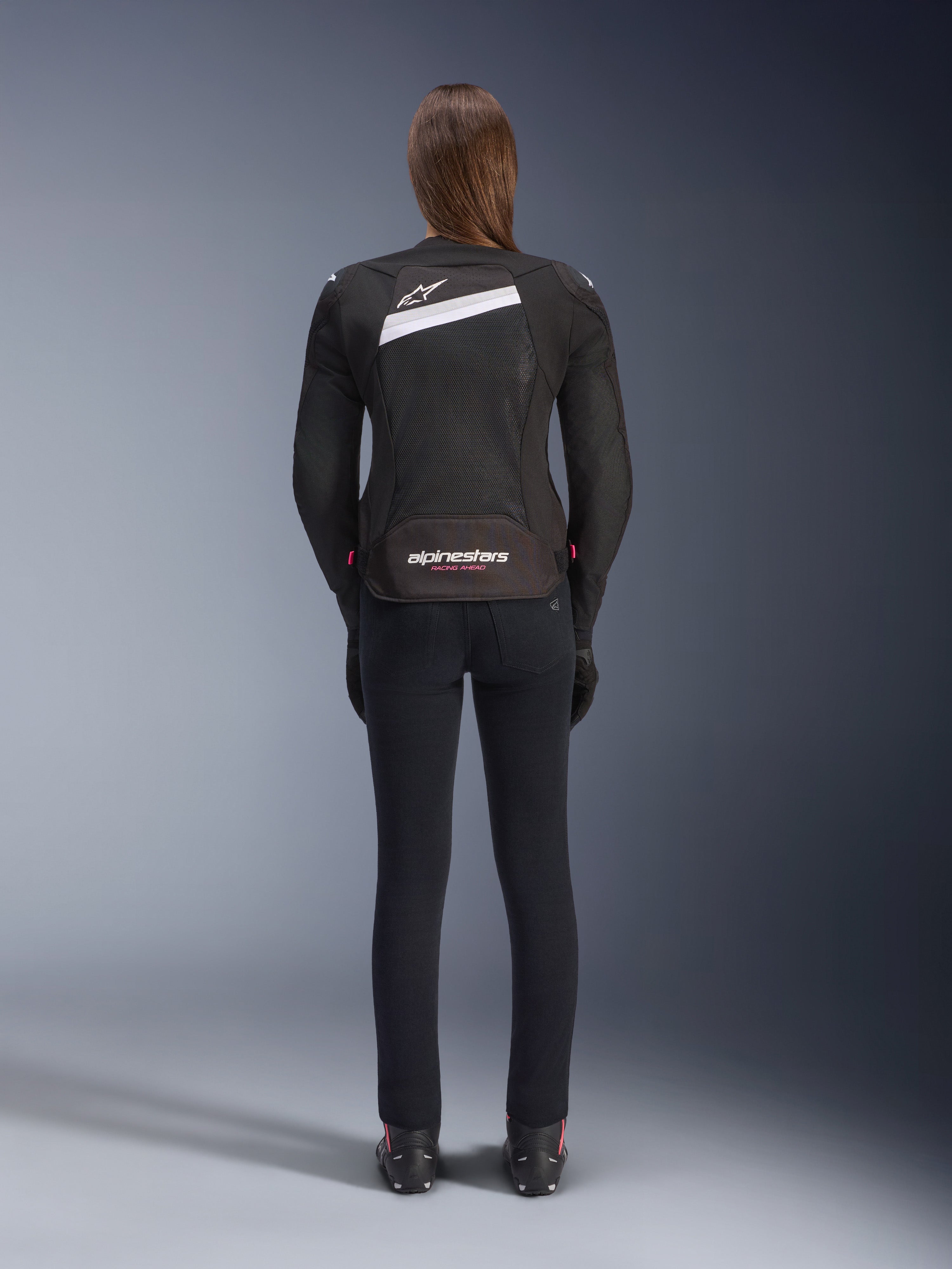 Giacca Alpinestars Stella T-GP Plus R V4 Airflow, nero e bianco, vista posteriore con ampi pannelli di ventilazione in mesh e marchio Alpinestars, indossata da una pilota con pantaloni da moto neri slim-fit e stivali, abbigliamento tecnico sportivo in tessuto da donna