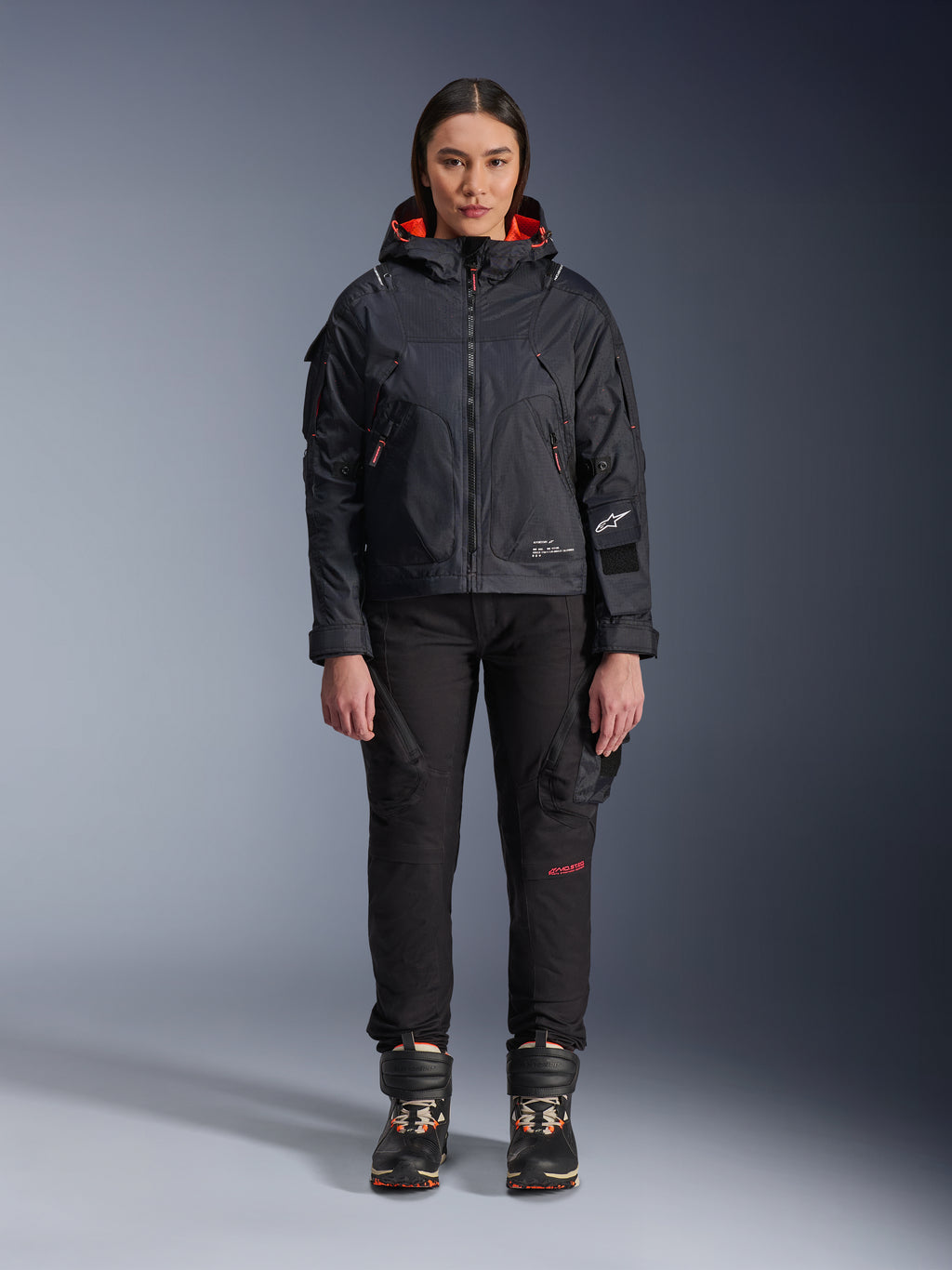 Modella che indossa la giacca Alpinestars Molly Air in nero e corallo fluo, caratterizzata da un design d'ispirazione utility con cappuccio e fodera interna corallo, abbinata a pantaloni da guida neri e stivali da moto, abbigliamento da moto urban contemporary
