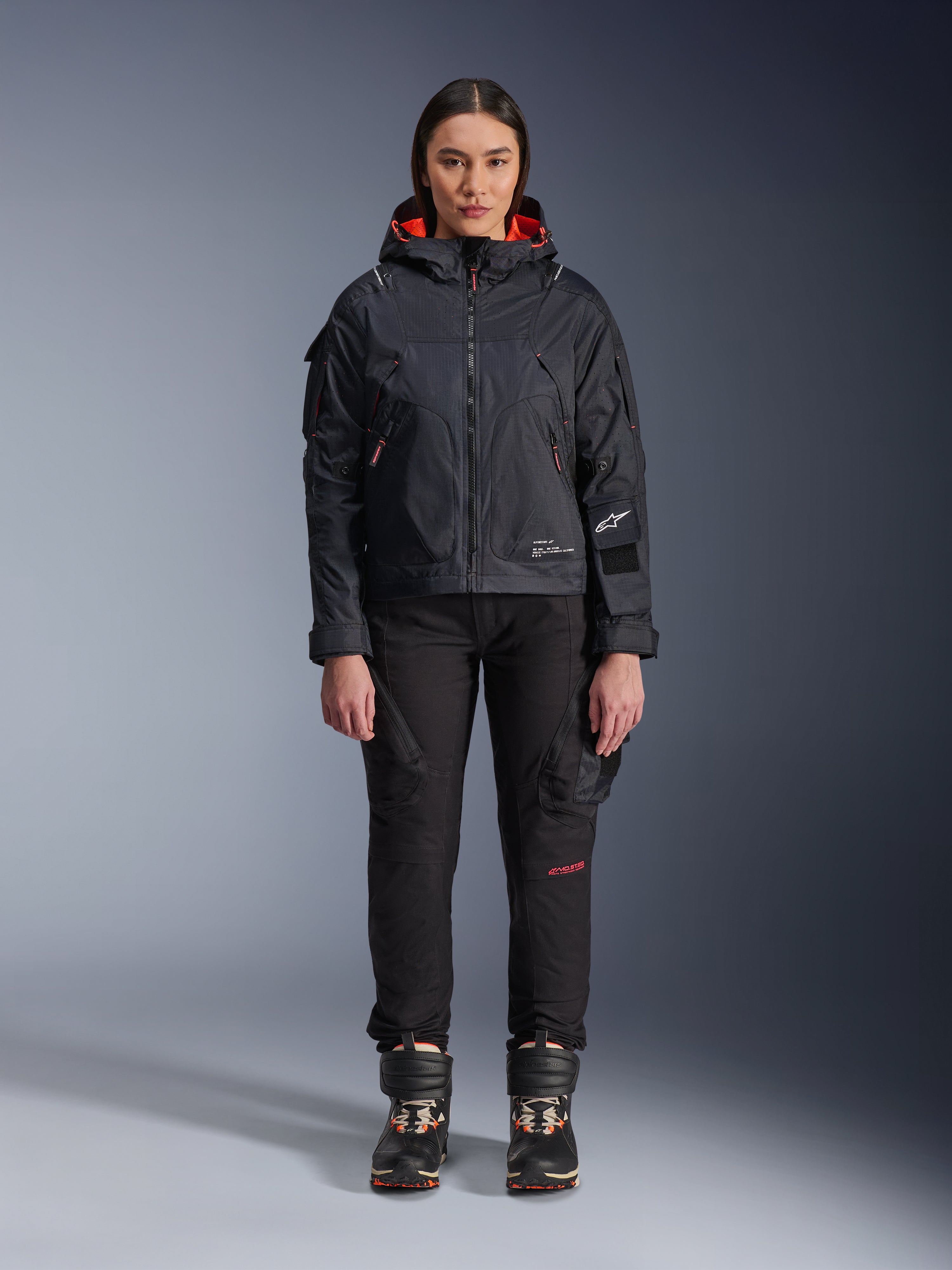 Modella che indossa la giacca Alpinestars Molly Air in nero e corallo fluo, caratterizzata da un design d'ispirazione utility con cappuccio e fodera interna corallo, abbinata a pantaloni da guida neri e stivali da moto, abbigliamento da moto urban contemporary