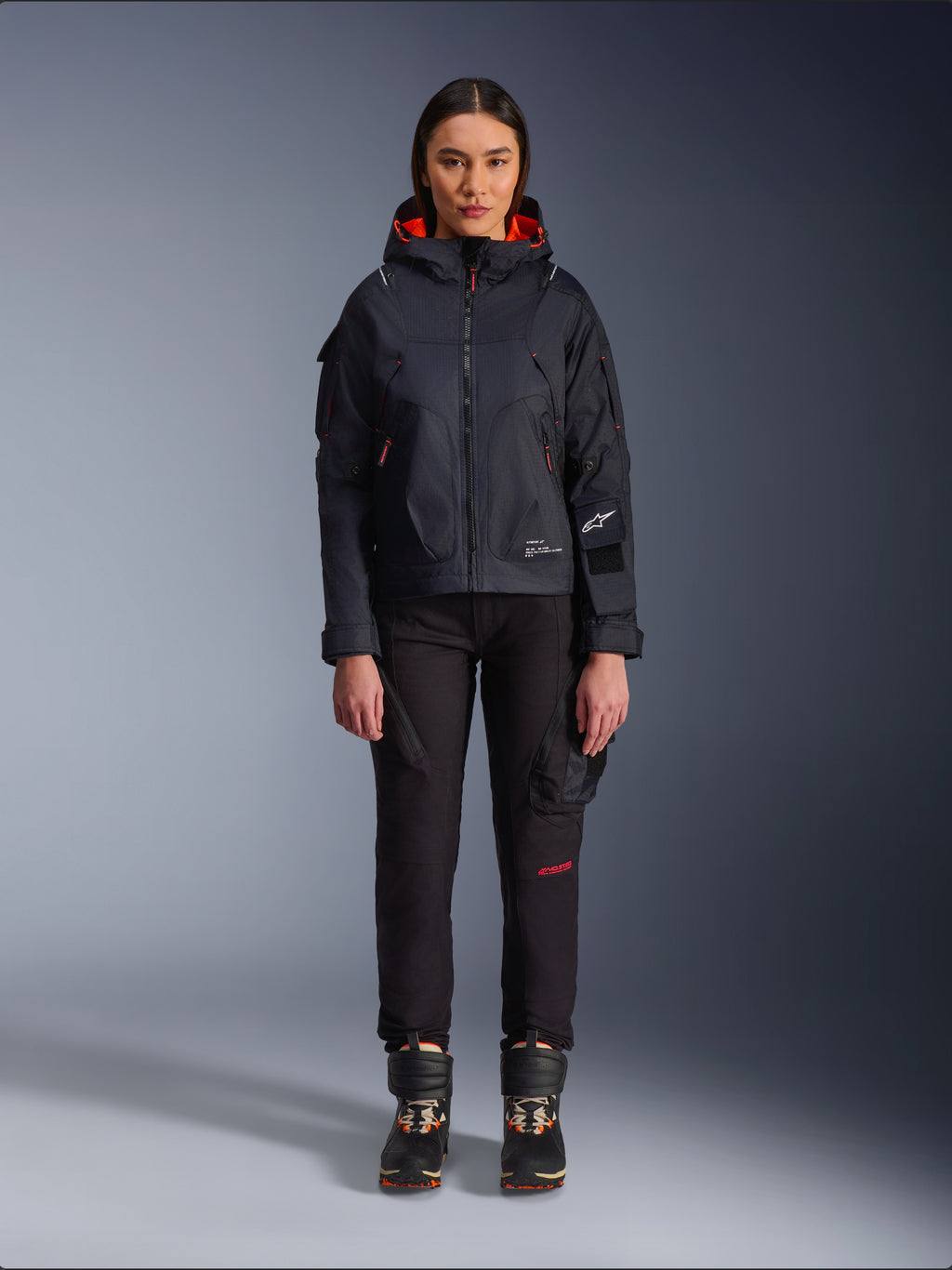 Pantaloni da donna Alpinestars Actea, pantaloni da moto neri con scritte rosse e tasche cargo, indossati da una motociclista con giacca nera con cappuccio, abbigliamento moto urban moderno