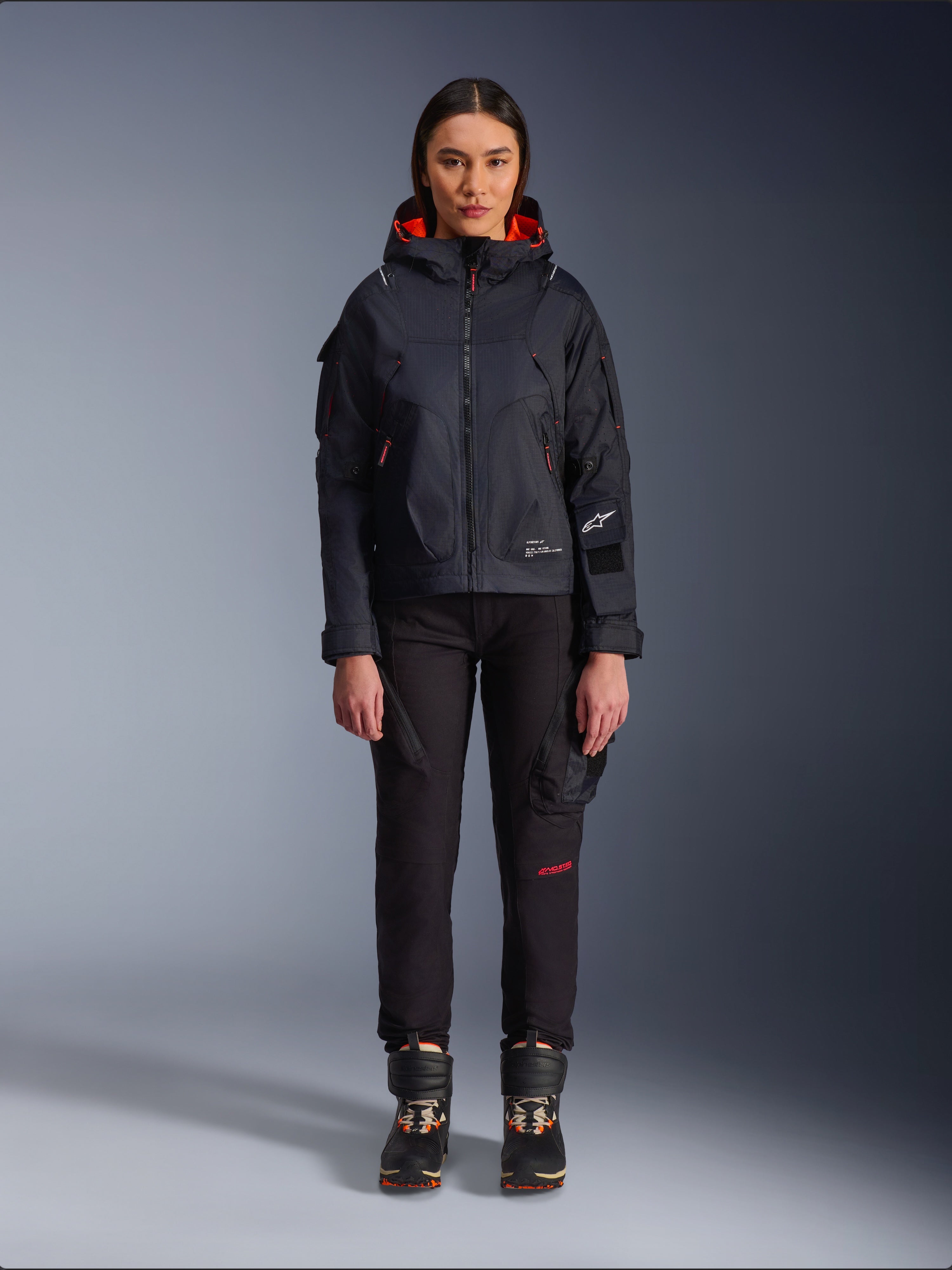 Pantaloni da donna Alpinestars Actea, pantaloni da moto neri con scritte rosse e tasche cargo, indossati da una motociclista con giacca nera con cappuccio, abbigliamento moto urban moderno