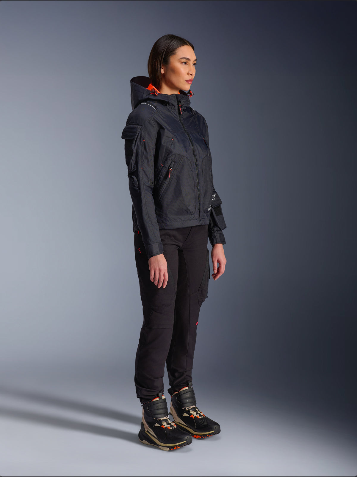 Motociclista che indossa la giacca Alpinestars Molly Air in Black Fluo Coral, giacca da moto in tessuto leggero con fodera del cappuccio arancione acceso, abbinata a pantaloni da guida neri e stivali tecnici, design d'ispirazione utility in stile urbano contemporaneo.