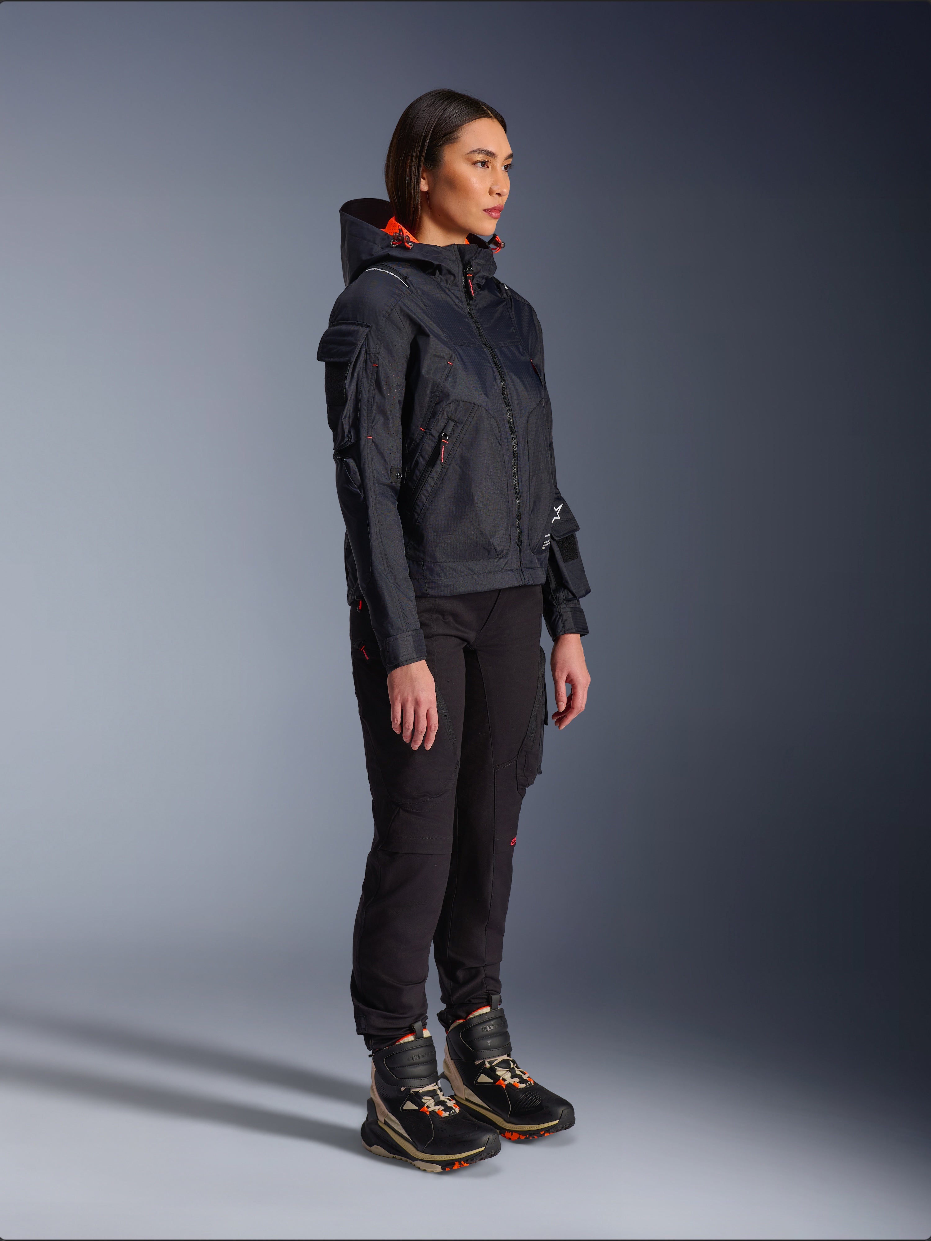 Alpinestars Pantaloni Actea da Donna in nero, vestibilità regolare con tasche cargo, abbinati a una giacca Molly Air nera con dettagli arancioni e scarpe da moto alte, abbigliamento motociclistico urbano contemporaneo per donna